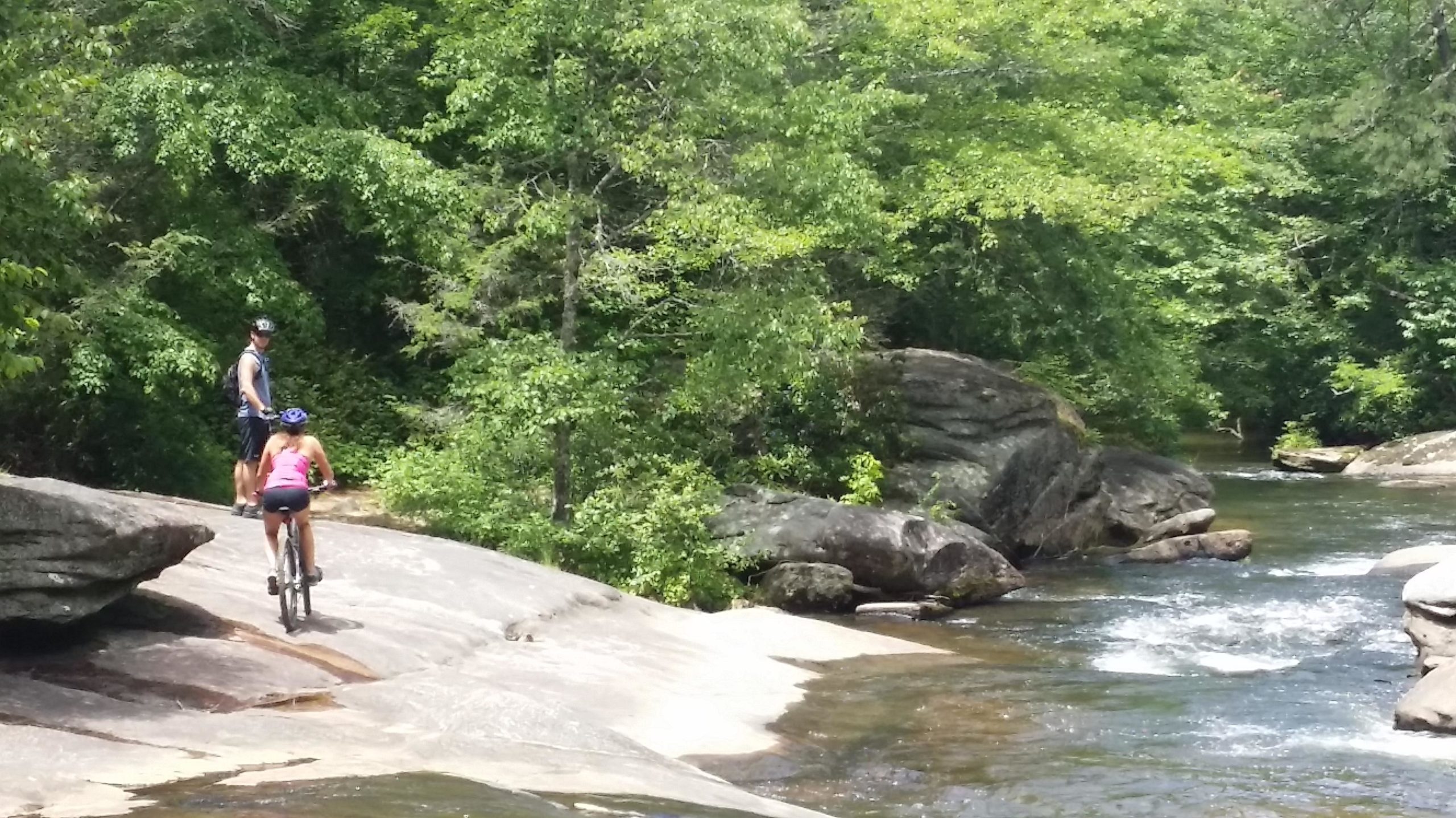Two people enjoy a sunny day outdoors, one riding a bicycle on a rocky path near a river, while the other stands nearby, surrounded by lush green trees and rocks. DuPont State Forest mountain bike trail.