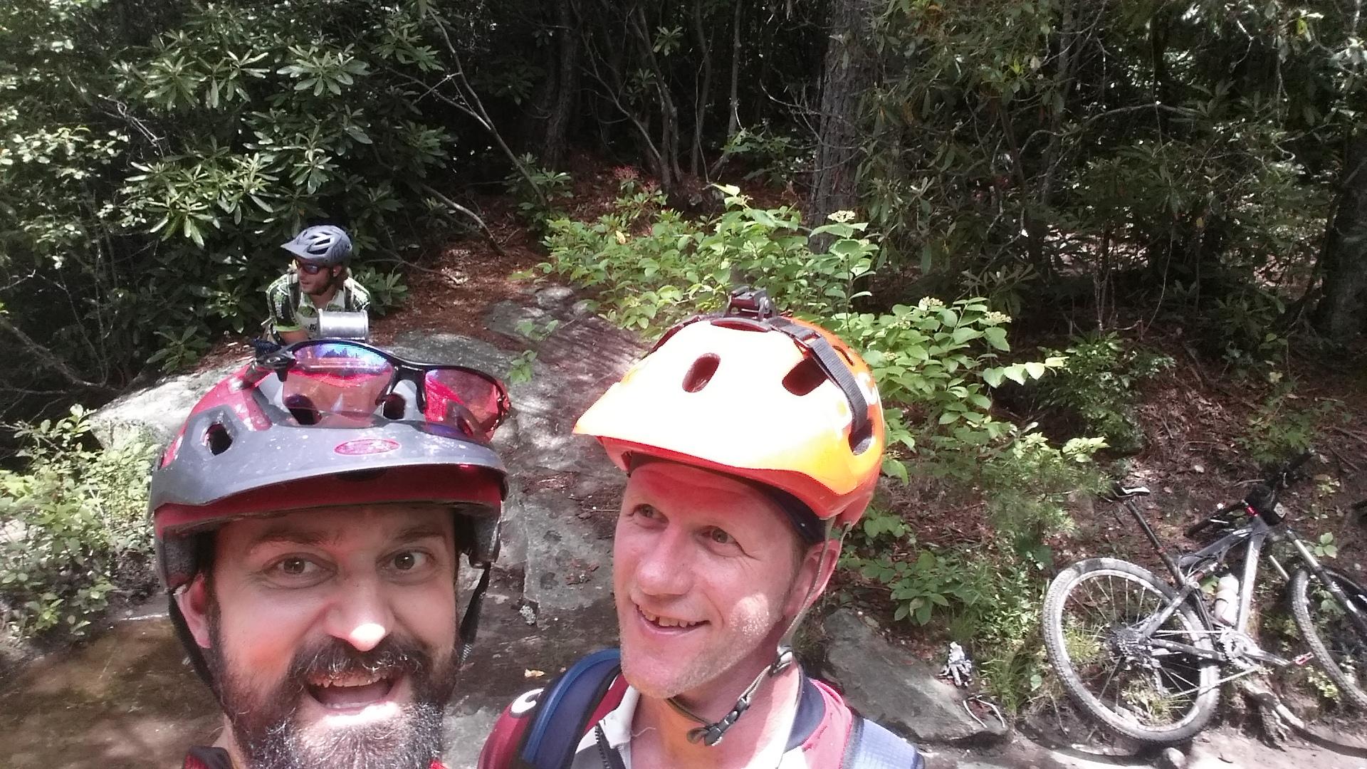 Two cyclists wearing helmets smile for a selfie on a wooded trail, surrounded by lush greenery. In the background, a third cyclist sits on a rock, partially visible, while a mountain bike rests nearby. DuPont State Recreational Forest mountain bike trail.