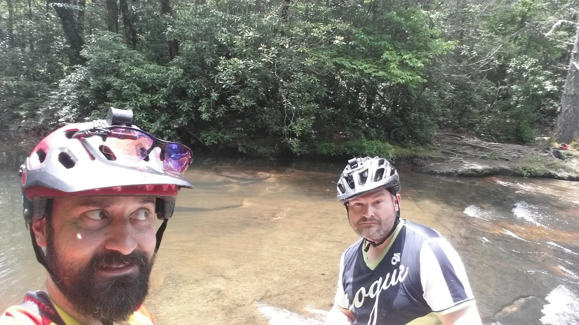 Two mountain bikers posing for a selfie at a shallow stream in a forested area. One biker wears a red helmet with a camera attached, while the other has a black and green cycling jersey. Lush green trees provide a backdrop, and the water appears clear and calm. DuPont State Forest mountain bike trail.