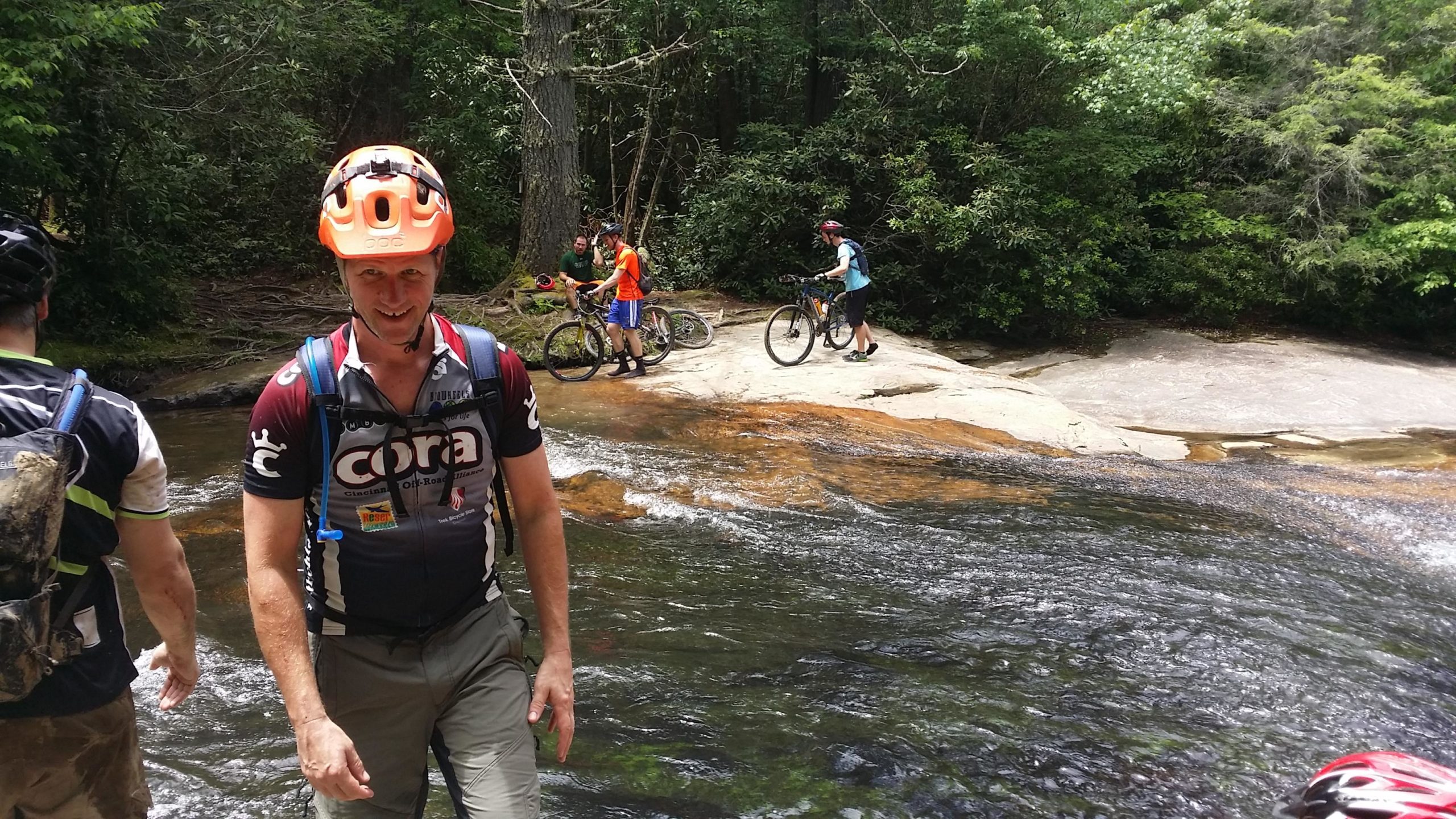 A group of mountain bikers near a stream in a wooded area. One biker in the foreground wears an orange helmet and a cycling jersey, smiling as he walks along the water. In the background, two individuals are seen with their bikes while another rider adjusts their equipment on the rocky bank. Lush greenery surrounds the scene, highlighting a sunny day outdoors. DuPont State Forest mountain bike trail.