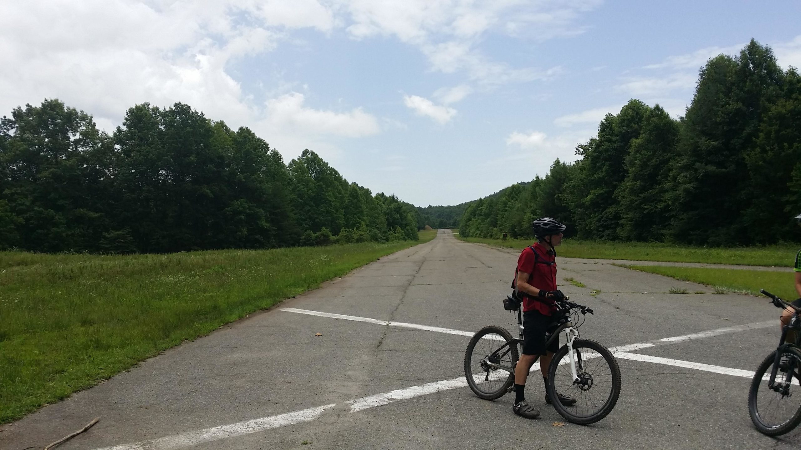 Two cyclists on mountain bikes stand near a large, overgrown, abandoned airstrip, surrounded by lush green trees and grass. The sky is partly cloudy, and the asphalt runway stretches into the distance. The cyclists are wearing helmets and casual riding gear, seemingly engaged in conversation while taking a break. DuPont State Forest mountain bike trail.