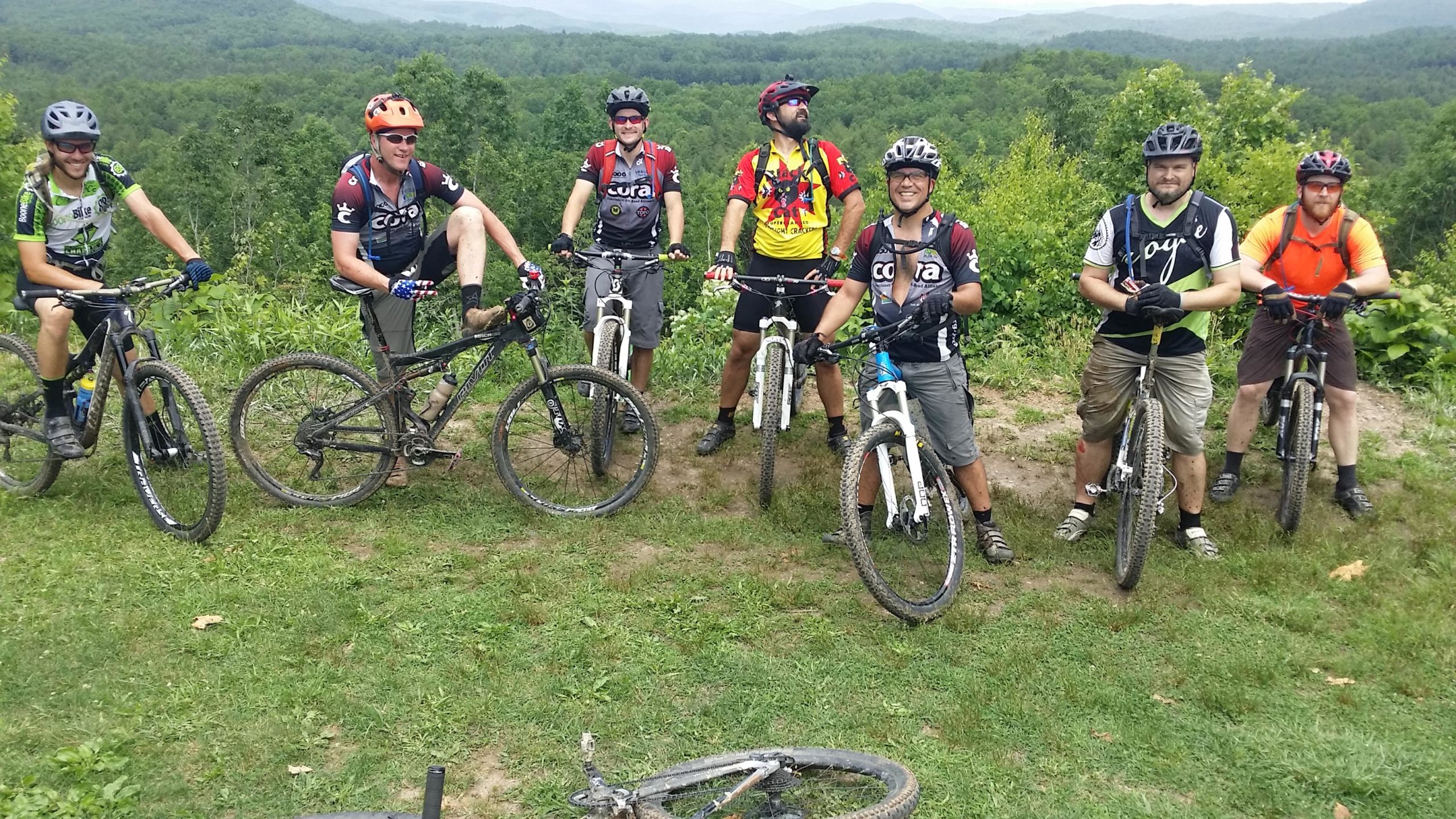 A group of seven mountain bikers poses for a photo on a grassy hilltop, showcasing their bikes. They are wearing cycling gear and helmets, with a scenic view of green mountains in the background. The cyclists appear happy and adventurous, with some of them displaying playful poses. The ground is slightly muddy, indicating they've been riding on trails. DuPont State Recreational Forest mountain bike trail.