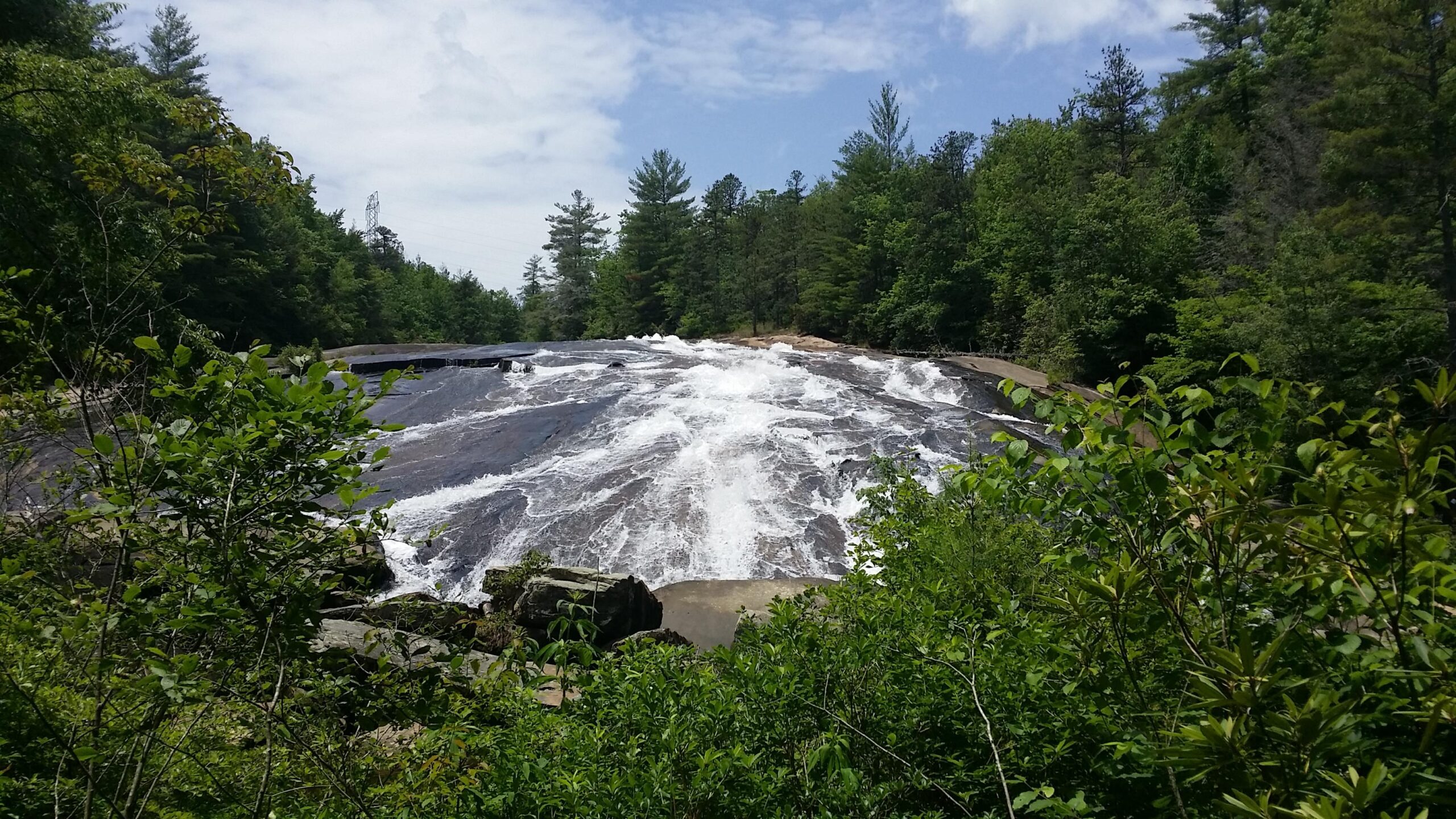 A scenic view of a waterfall cascading over a rocky surface, surrounded by lush green trees and foliage under a partly cloudy sky. The water flows energetically, creating white frothy waves as it descends. DuPont State Recreational Forest mountain bike trail.