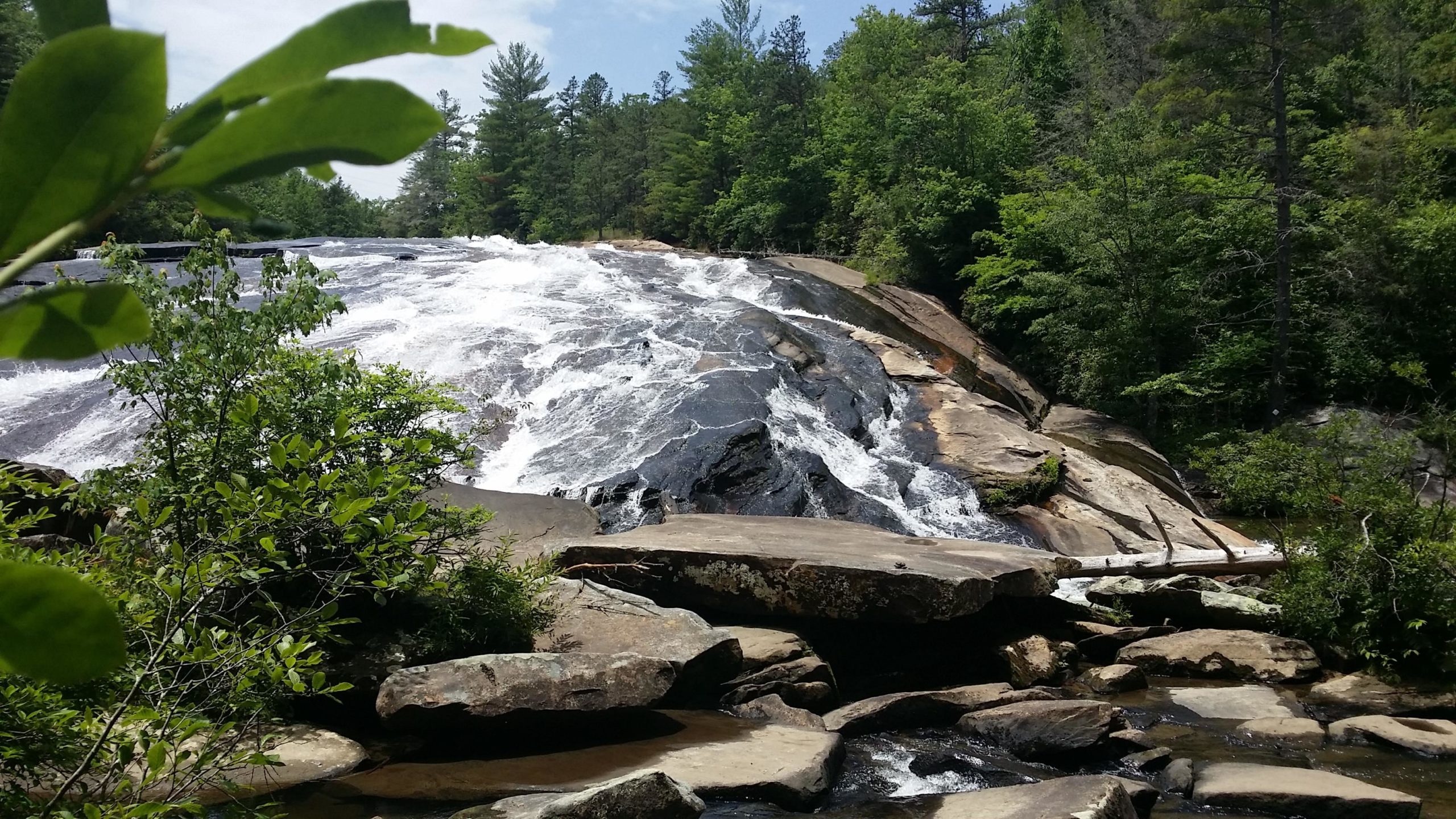 A cascading waterfall flowing over rocky terrain, surrounded by lush green trees and vegetation under a bright blue sky. The water creates a frothy white spray as it descends, highlighting the natural beauty of the landscape. DuPont State Recreational Forest mountain bike trail.