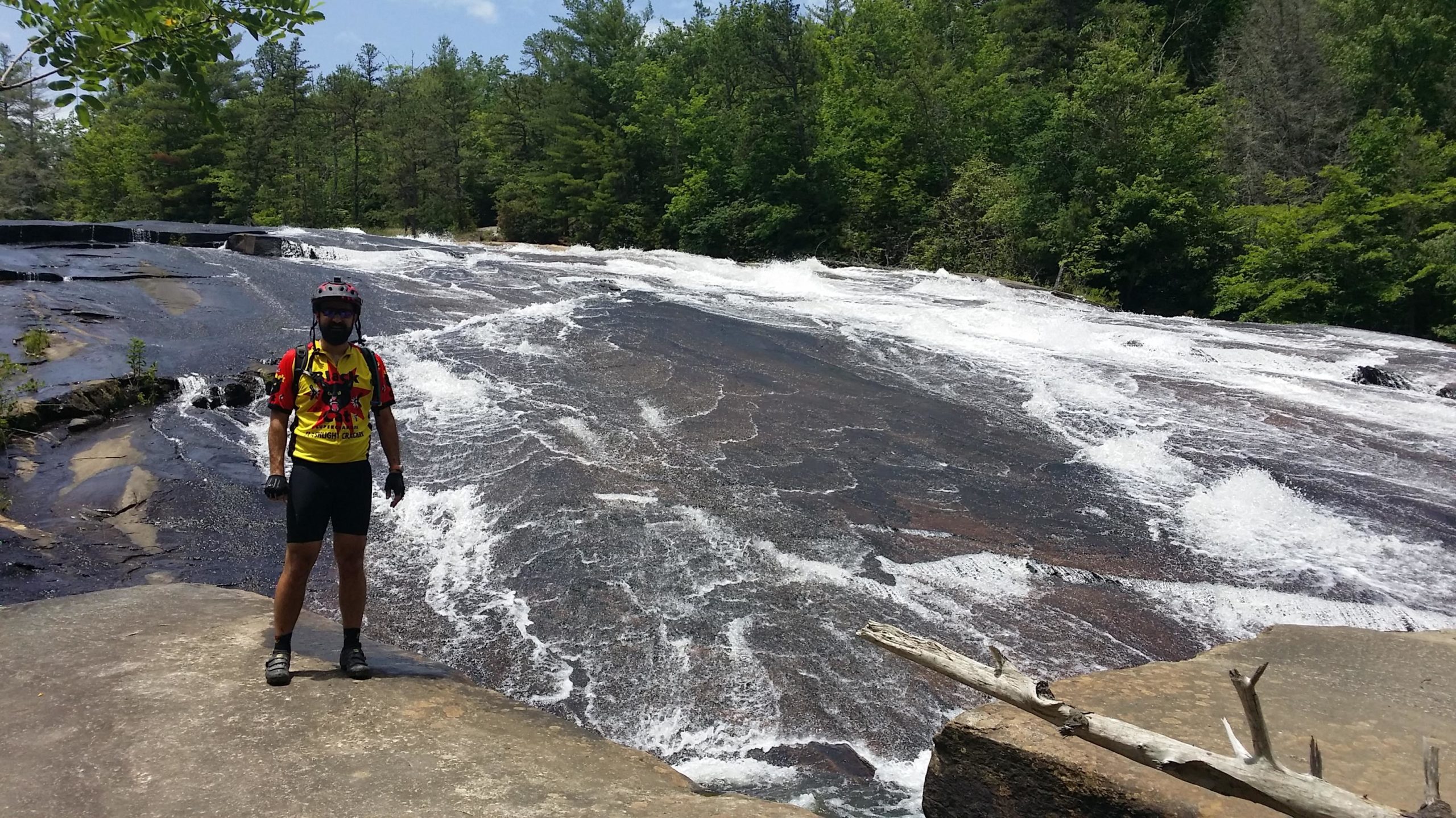 A cyclist standing on a rocky outcrop next to a flowing waterfall. The cyclist is wearing a bright yellow and red jersey, with a helmet and gloves, and is surrounded by lush green trees in the background. The water cascades over smooth rocks, creating foamy white rapids. DuPont State Forest mountain bike trail.