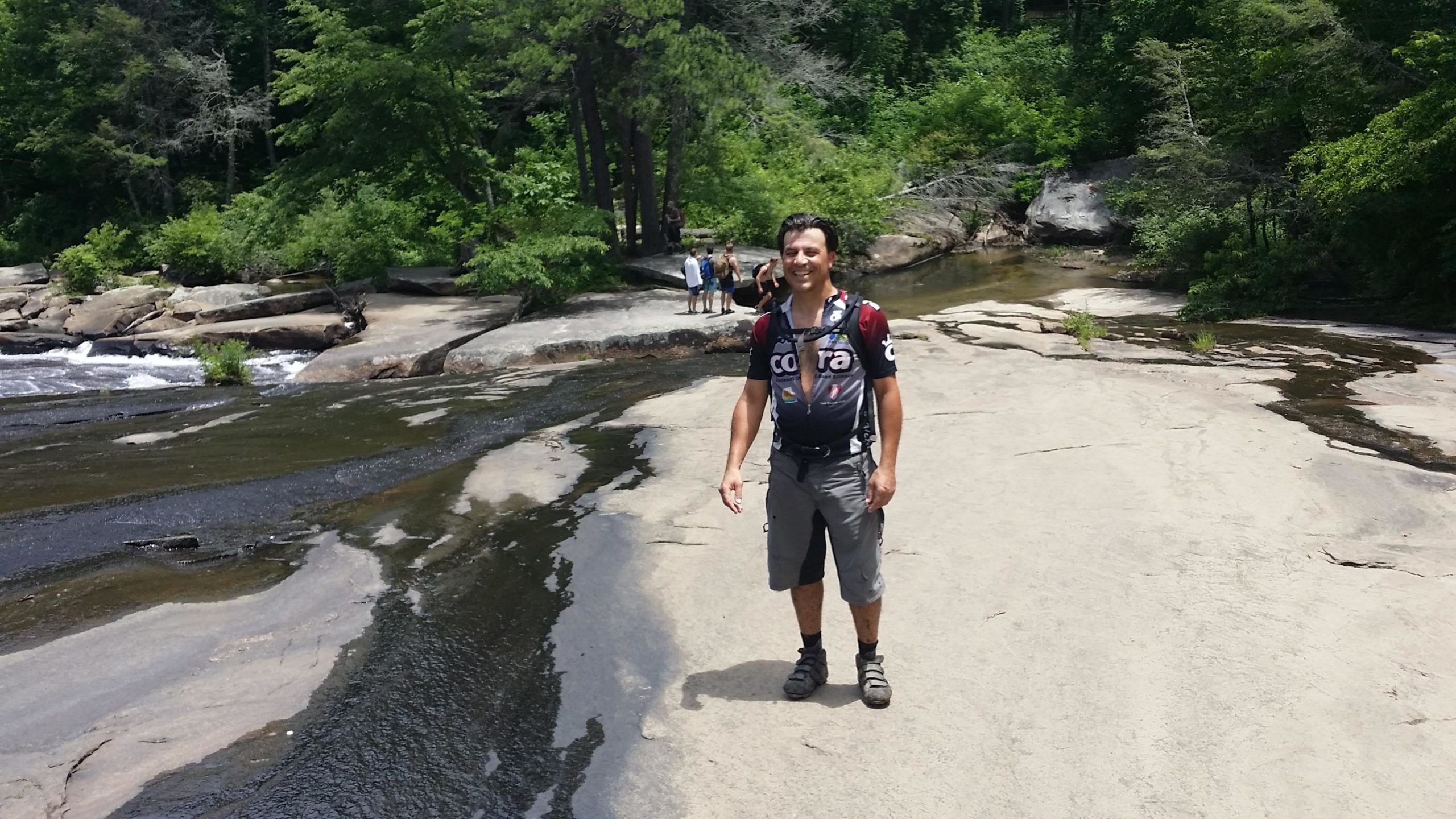 A person standing on smooth, rocky terrain near a flowing stream, surrounded by lush greenery and trees. In the background, a few people can be seen, enjoying the natural setting. The individual is wearing a sports shirt, shorts, and sandals, with a friendly expression. DuPont State Recreational Forest mountain bike trail.