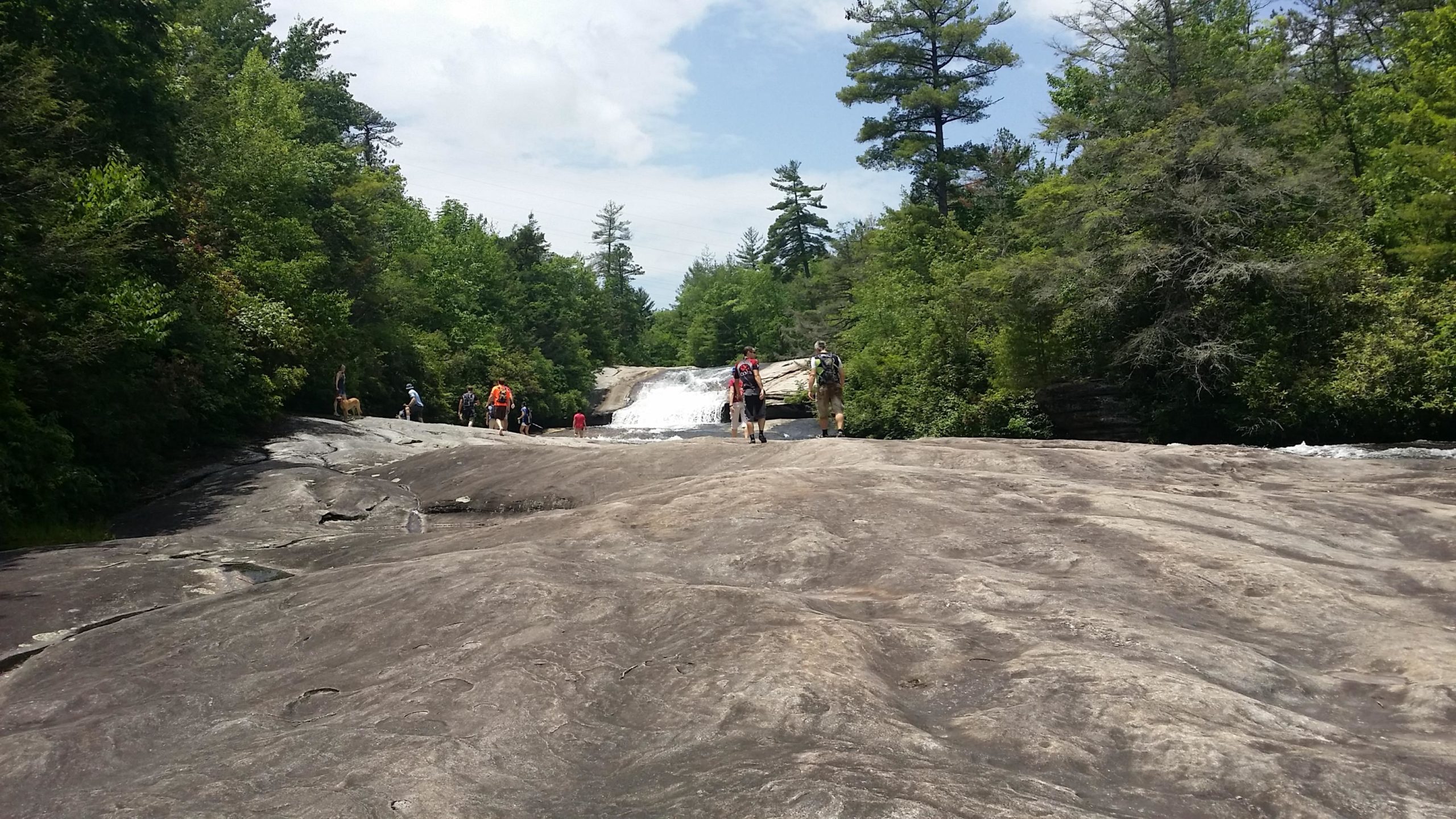A group of hikers explores a rocky surface near a waterfall, surrounded by lush greenery and trees under a cloudy sky. DuPont State Recreational Forest mountain bike trail.