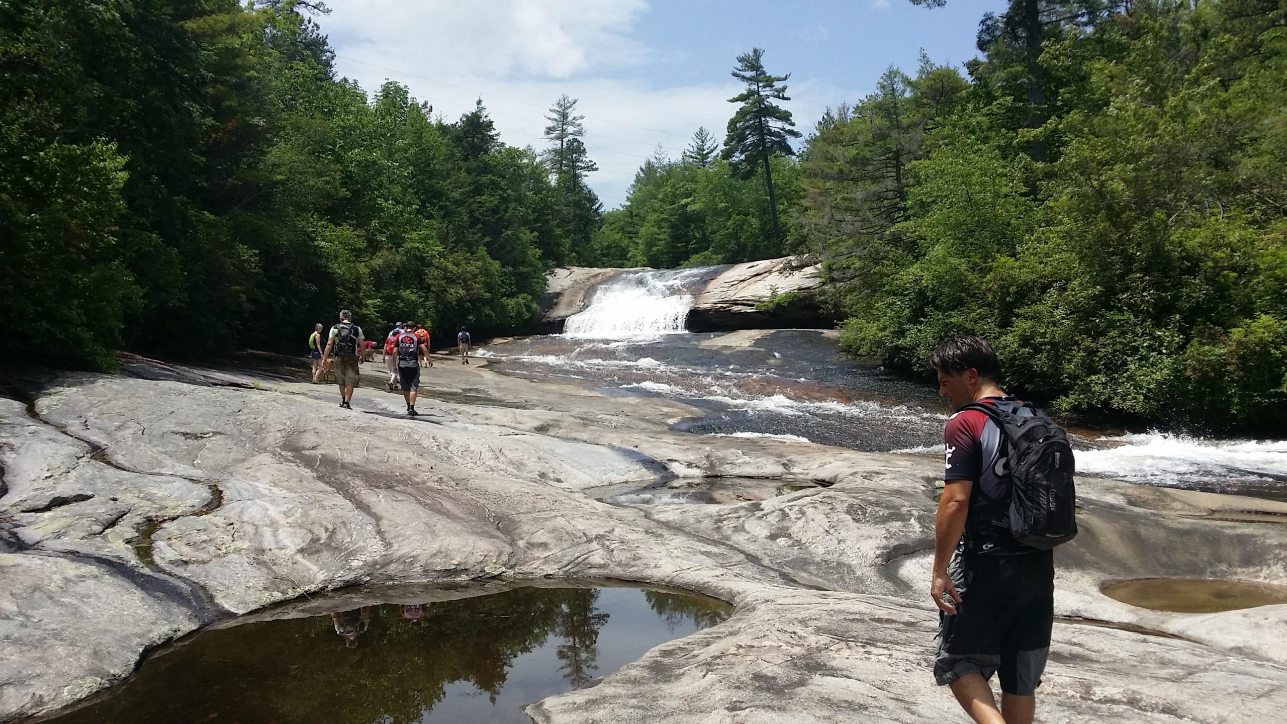 A scenic view of a waterfall cascading down smooth rocks, surrounded by lush green trees. In the foreground, a group of hikers are walking along the rocky terrain, while one person stands closer to the water's edge, observing the surroundings. The sky is partly cloudy, and the natural beauty of the area is highlighted. DuPont State Recreational Forest mountain bike trail.