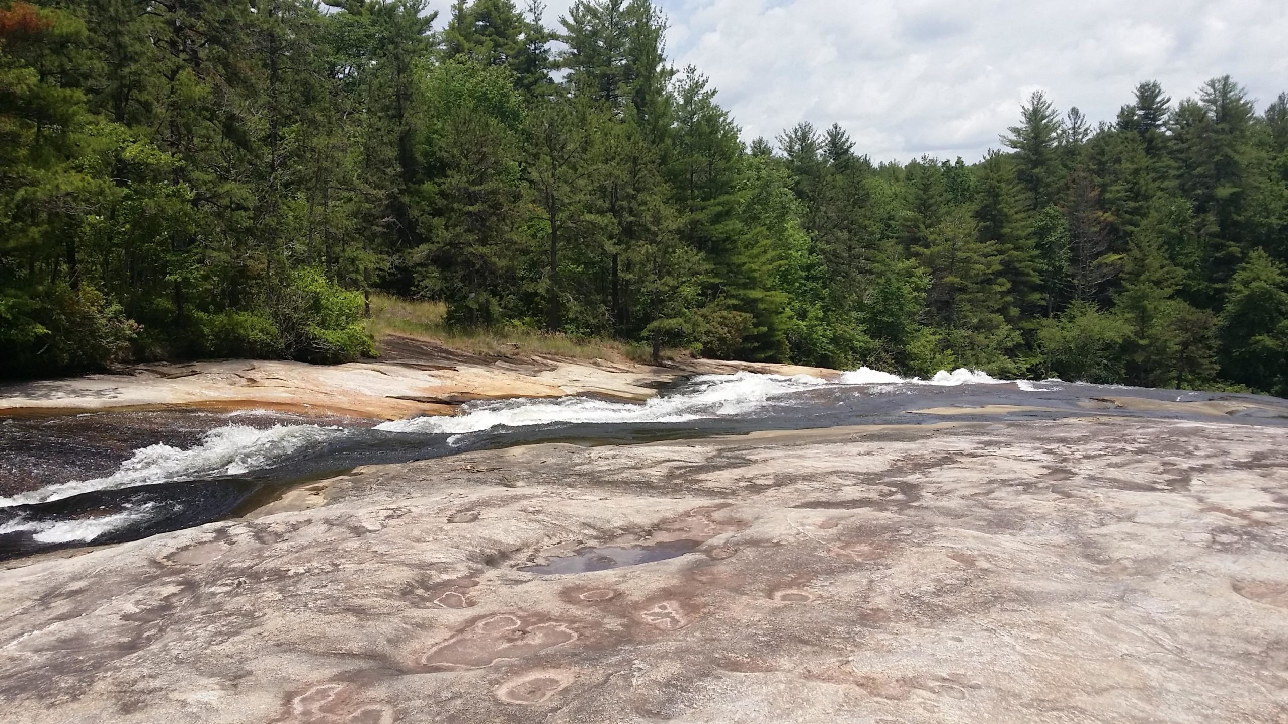 A scenic view of a river flowing over smooth, rocky terrain surrounded by lush green trees under a partly cloudy sky. DuPont State Recreational Forest mountain bike trail.