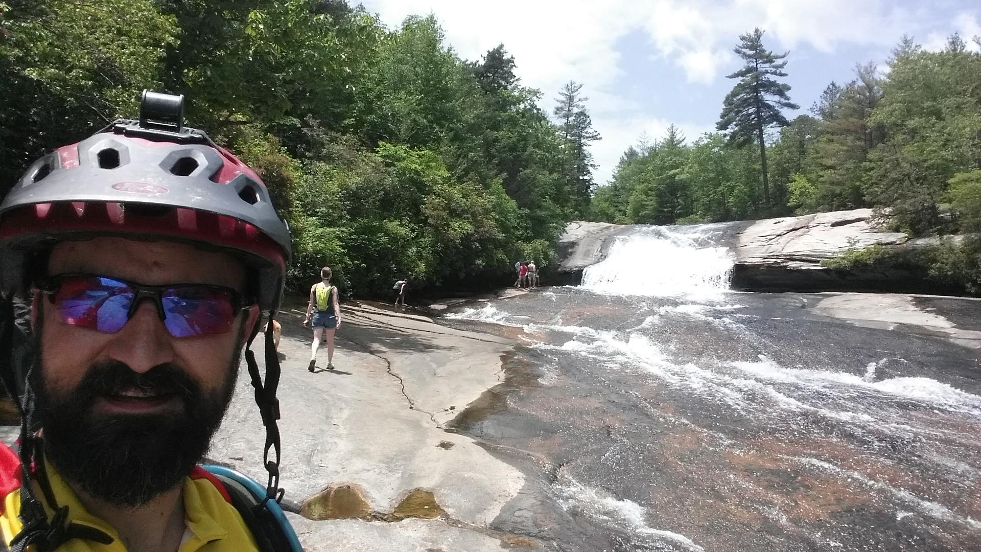 A person wearing a biking helmet and sunglasses poses for a selfie in the foreground, with a waterfall and rocky terrain in the background. Several individuals can be seen exploring the area near the waterfall, surrounded by lush green trees and a bright blue sky. DuPont State Forest mountain bike trail.