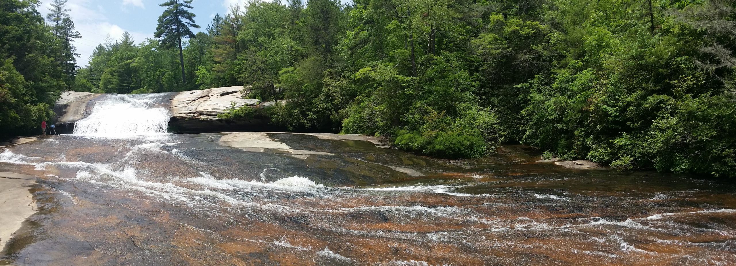 A scenic view of a waterfall cascading over smooth, rocky terrain, surrounded by lush greenery and trees. The water flows over a wide, sloped rock surface, creating a gentle rush. A few people can be seen enjoying the natural setting near the water's edge under a clear blue sky. DuPont State Forest mountain bike trail.