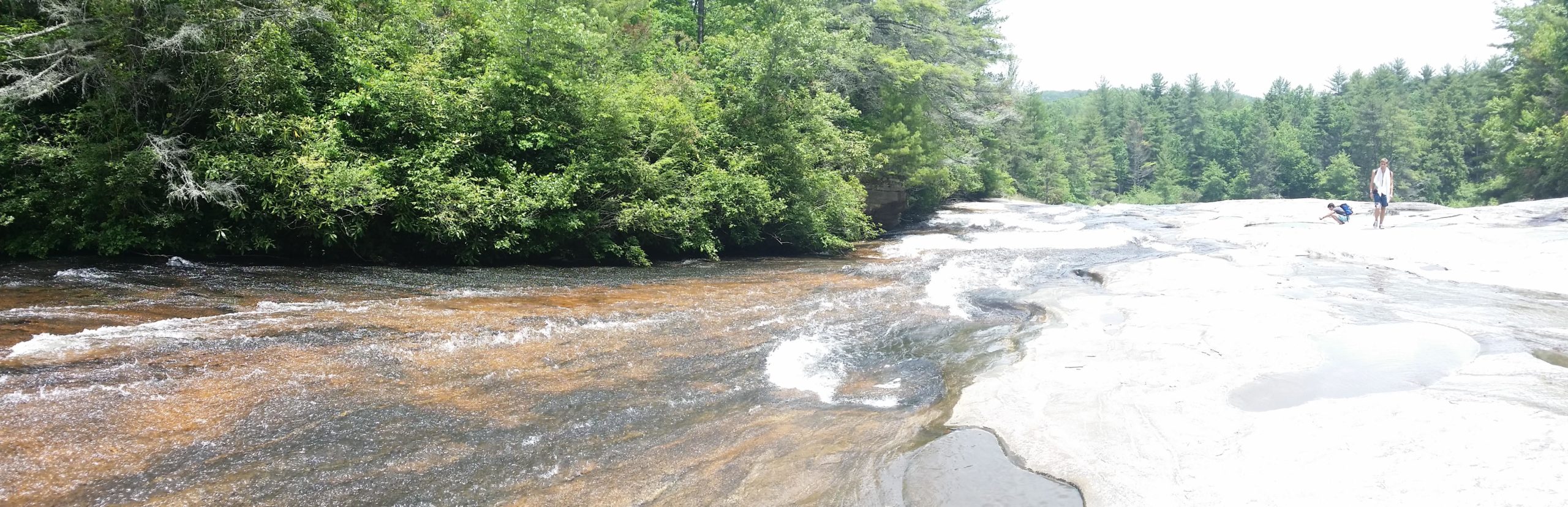 A scenic view of a rocky riverbed with flowing water surrounded by lush green trees. Two people are in the foreground; one is walking on the smooth rocks while another is kneeling near the water's edge. The sunlight creates a bright atmosphere, highlighting the natural beauty of the landscape. DuPont State Recreational Forest mountain bike trail.