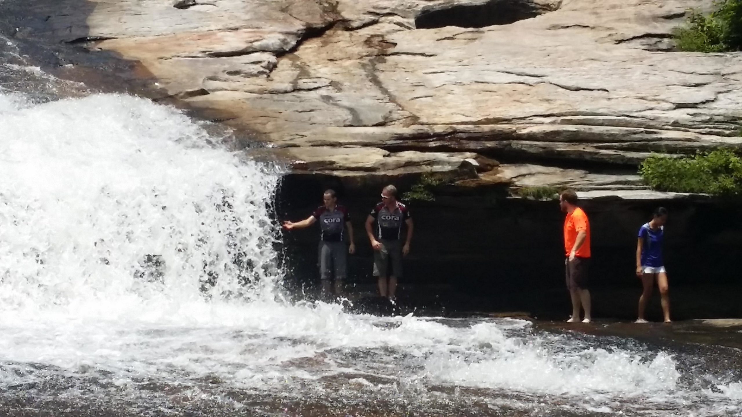 A group of four people standing near a waterfall in a rocky area. Two individuals are partially behind the cascading water, while the others are on the shore, enjoying the surroundings. The environment features natural rock formations and splashing water. They are dressed in casual outdoor clothing, with one person wearing an orange shirt and another in a blue shirt. DuPont State Forest mountain bike trail.