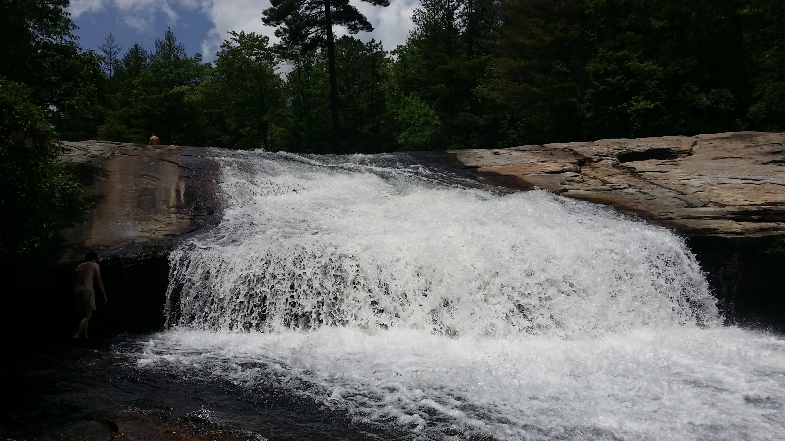 A scenic waterfall cascading over a rocky outcrop surrounded by lush green trees, with two individuals near the water's edge, enjoying the natural beauty on a sunny day. DuPont State Recreational Forest mountain bike trail.
