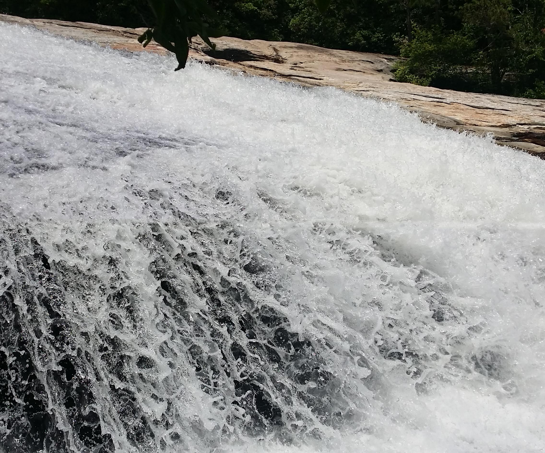 A close-up view of cascading water over smooth rocks, creating a frothy surface as it flows downward. The scene is surrounded by lush greenery in the background, highlighting the natural beauty of the waterfall. DuPont State Forest mountain bike trail.