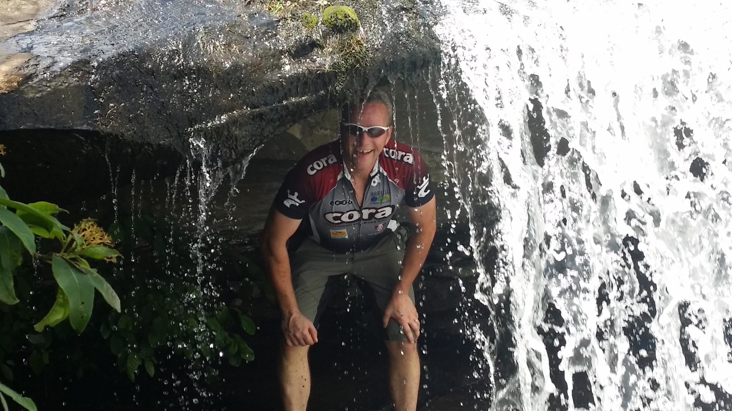 A person in a cycling jersey poses playfully under a waterfall, with water cascading over a rock formation. Green foliage is visible in the foreground, adding to the natural setting. DuPont State Recreational Forest mountain bike trail.