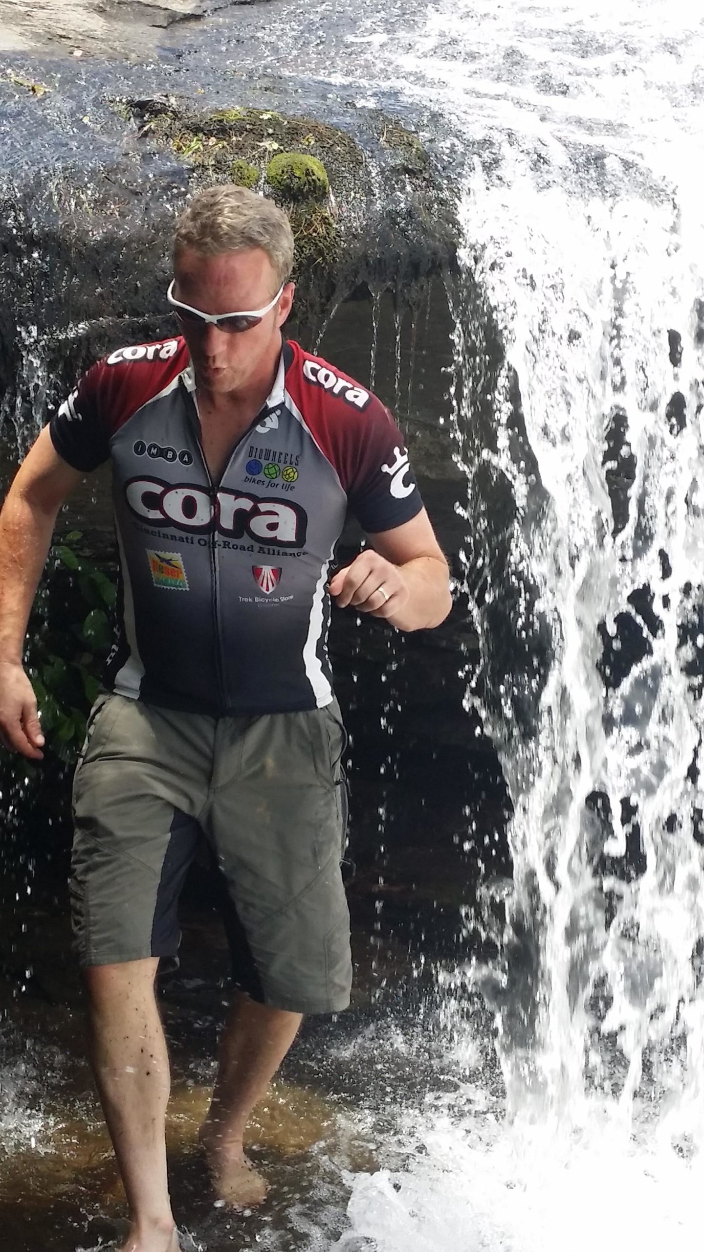 A person wearing sunglasses and a cycling jersey stands in shallow water at the base of a waterfall, with water cascading around them. They appear to be outdoors, enjoying a moment by the water. DuPont State Forest mountain bike trail.