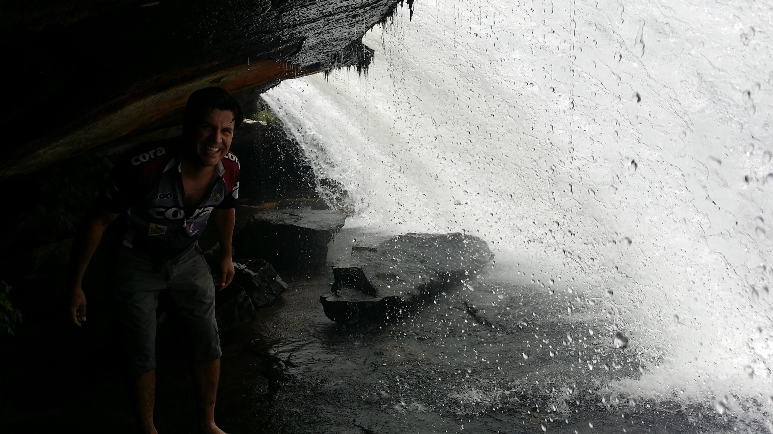A person standing under a rocky overhang near a waterfall, smiling and posing as water cascades down in the background, creating a misty spray. The scene captures the natural beauty of the waterfall and the person's enjoyment. DuPont State Forest mountain bike trail.