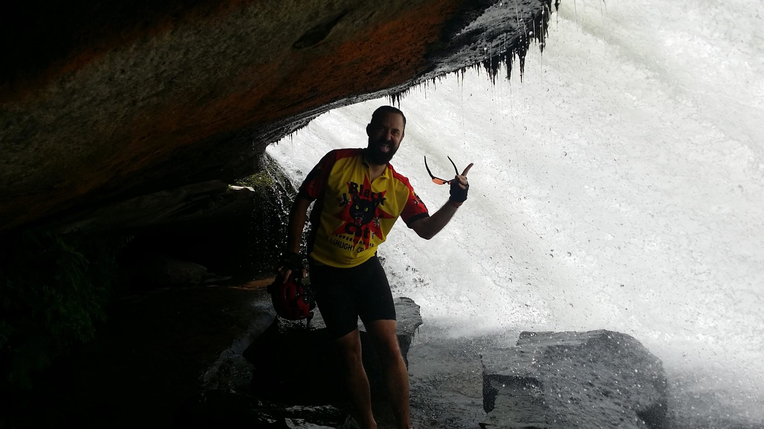 A person standing under a rock overhang near a waterfall, wearing a colorful cycling jersey and shorts. They are making a playful gesture and holding a helmet in one hand. Water splashes in the background, creating a misty atmosphere. DuPont State Recreational Forest mountain bike trail.
