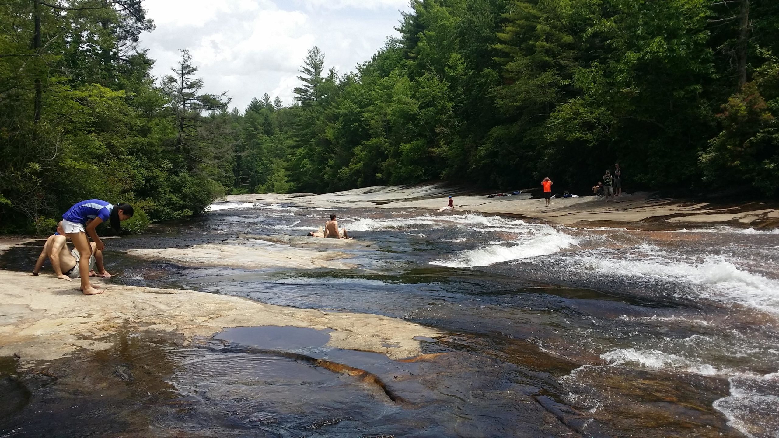 A scenic view of a rocky riverbank where several people are enjoying the water. The scene features a flowing river with gentle rapids, surrounded by lush green trees on both sides. Two individuals are playing near the water, while others are scattered along the river, some dipping their feet in the water. The sky is partly cloudy, providing a bright and inviting atmosphere. DuPont State Recreational Forest mountain bike trail.