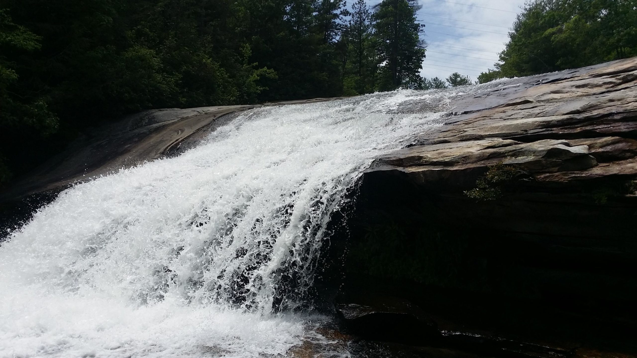 A cascading waterfall flowing over smooth, rocky terrain, surrounded by lush green trees under a partly cloudy sky. The water creates a white frothy spray as it descends, reflecting the natural beauty of the landscape. DuPont State Recreational Forest mountain bike trail.