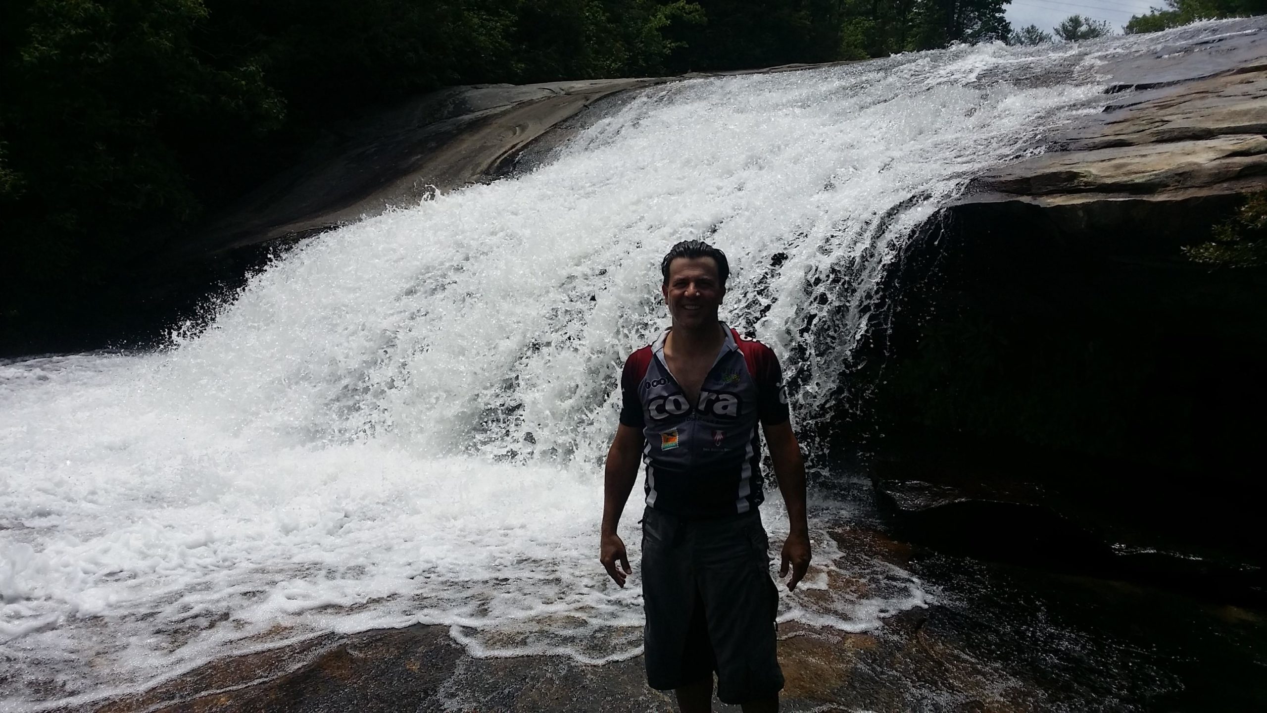 A person standing in front of a cascading waterfall, surrounded by greenery, with sunlight reflecting off the flowing water. The individual is smiling and wearing a cycling jersey and shorts, standing on a rocky surface in a wet environment. DuPont State Recreational Forest mountain bike trail.