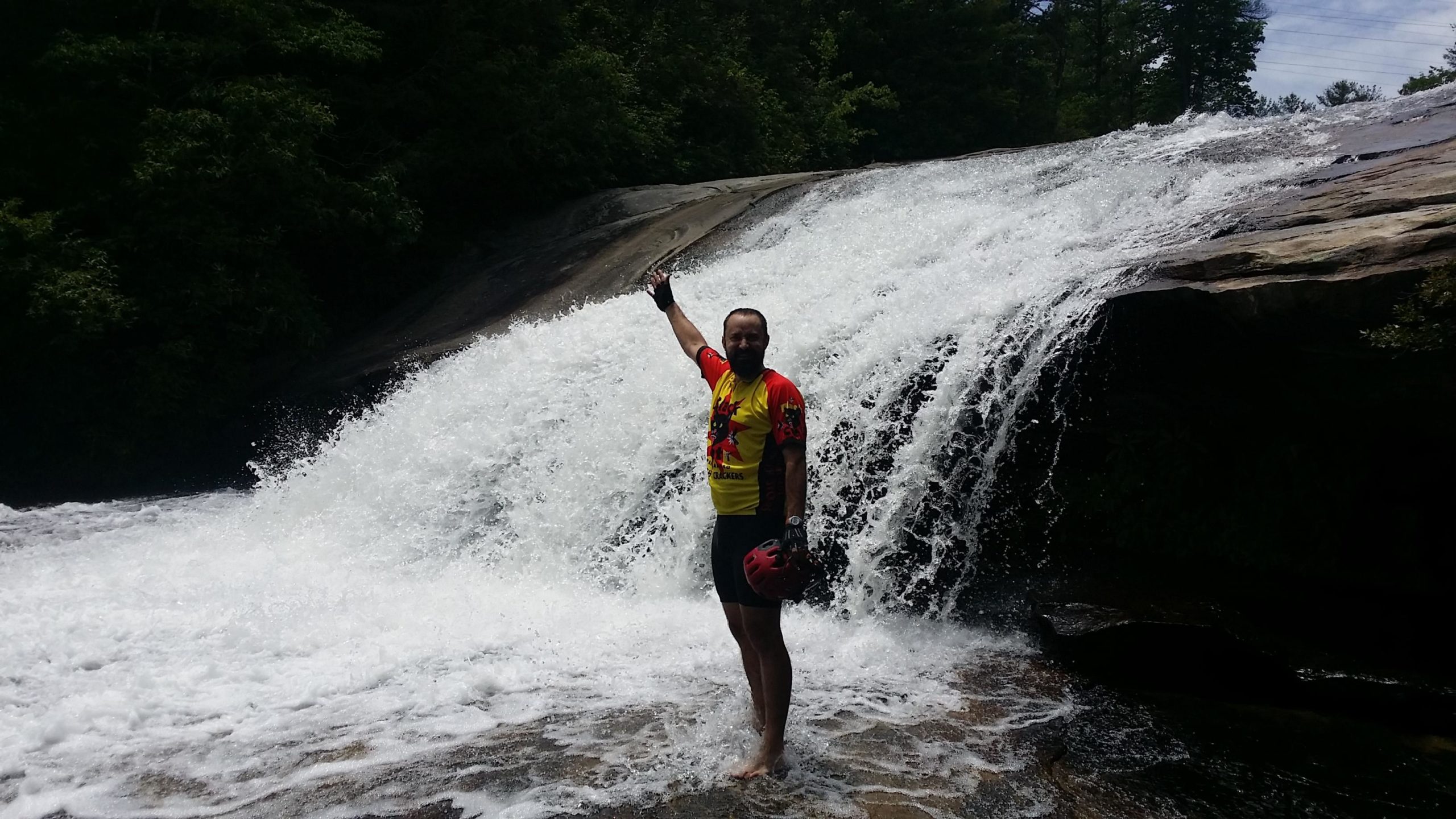 A person standing in shallow water at the base of a waterfall, raising one hand in celebration. They are wearing a bright yellow and red shirt and a helmet, with a rocky surface and lush greenery in the background. The waterfall cascades over a smooth rock surface, creating a splash of white water around them. DuPont State Forest mountain bike trail.