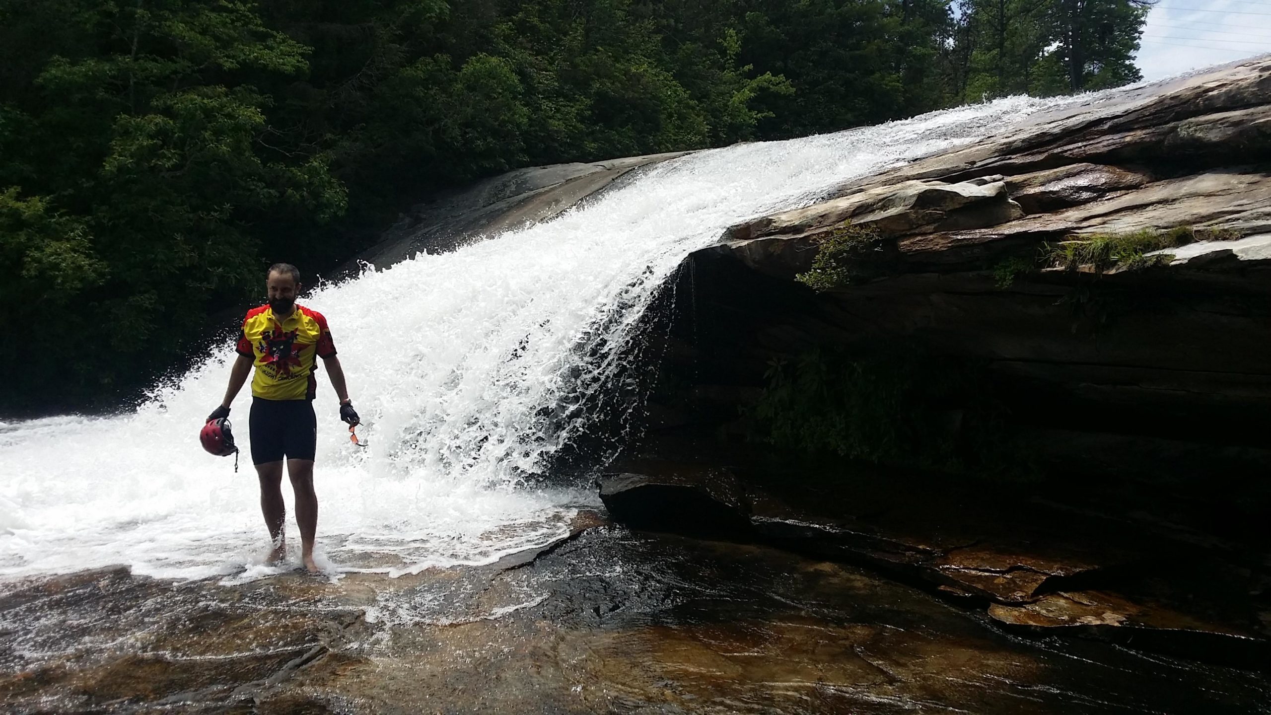 A person wearing a bright yellow shirt is standing in shallow water at the base of a waterfall, with water cascading down rocks in the background. The scene is surrounded by greenery, creating a natural and vibrant outdoor setting. The individual is holding a helmet and an object in one hand, while water splashes around their feet. DuPont State Forest mountain bike trail.