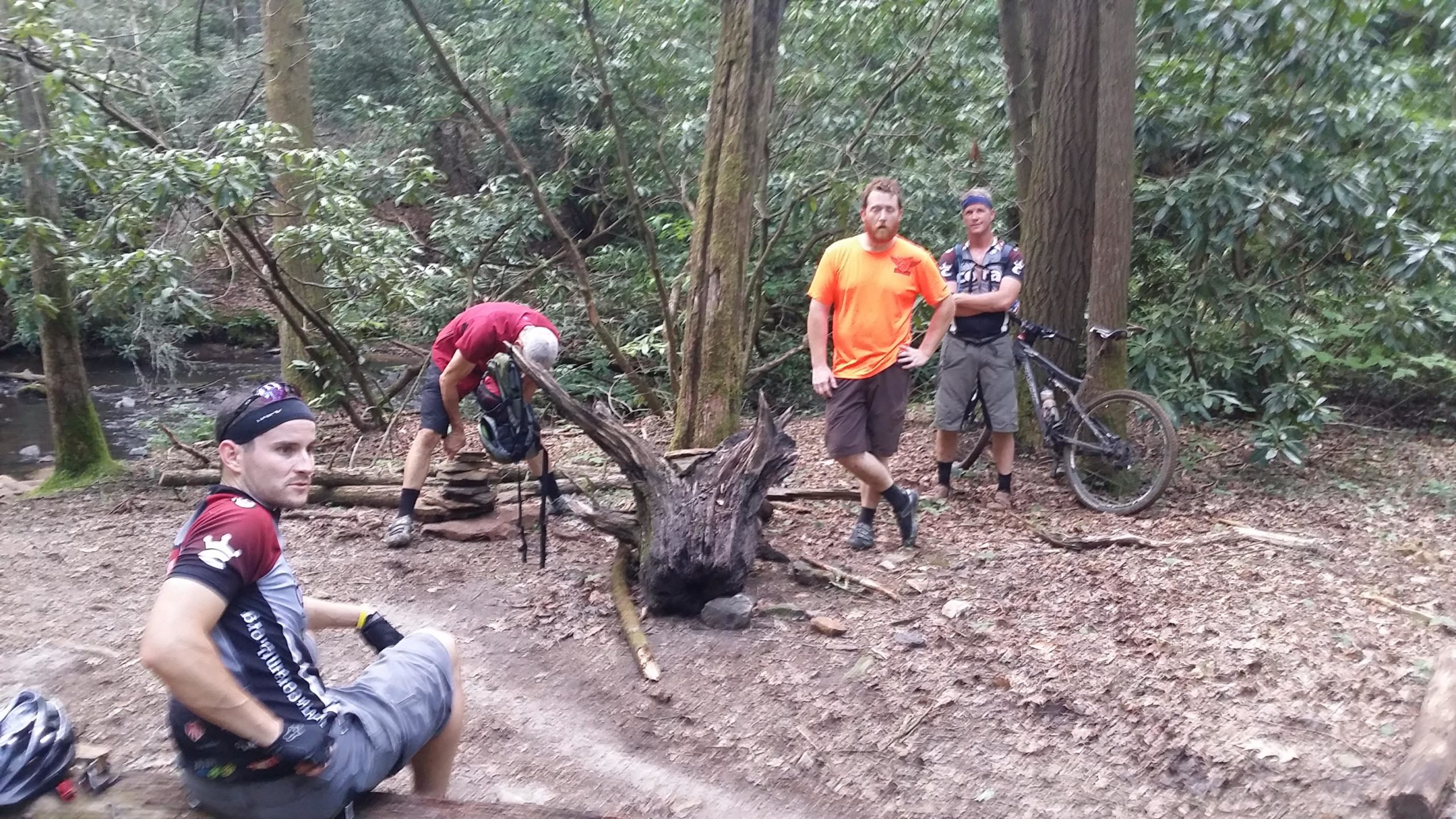 Four men are gathered in a wooded area near a stream, engaging in a break during a mountain biking trip. One man is sitting on a log, while the others are standing nearby, with one crouching to adjust his gear. They are surrounded by lush greenery and trees, with a mountain bike leaning against a tree in the background. DuPont State Forest mountain bike trail.