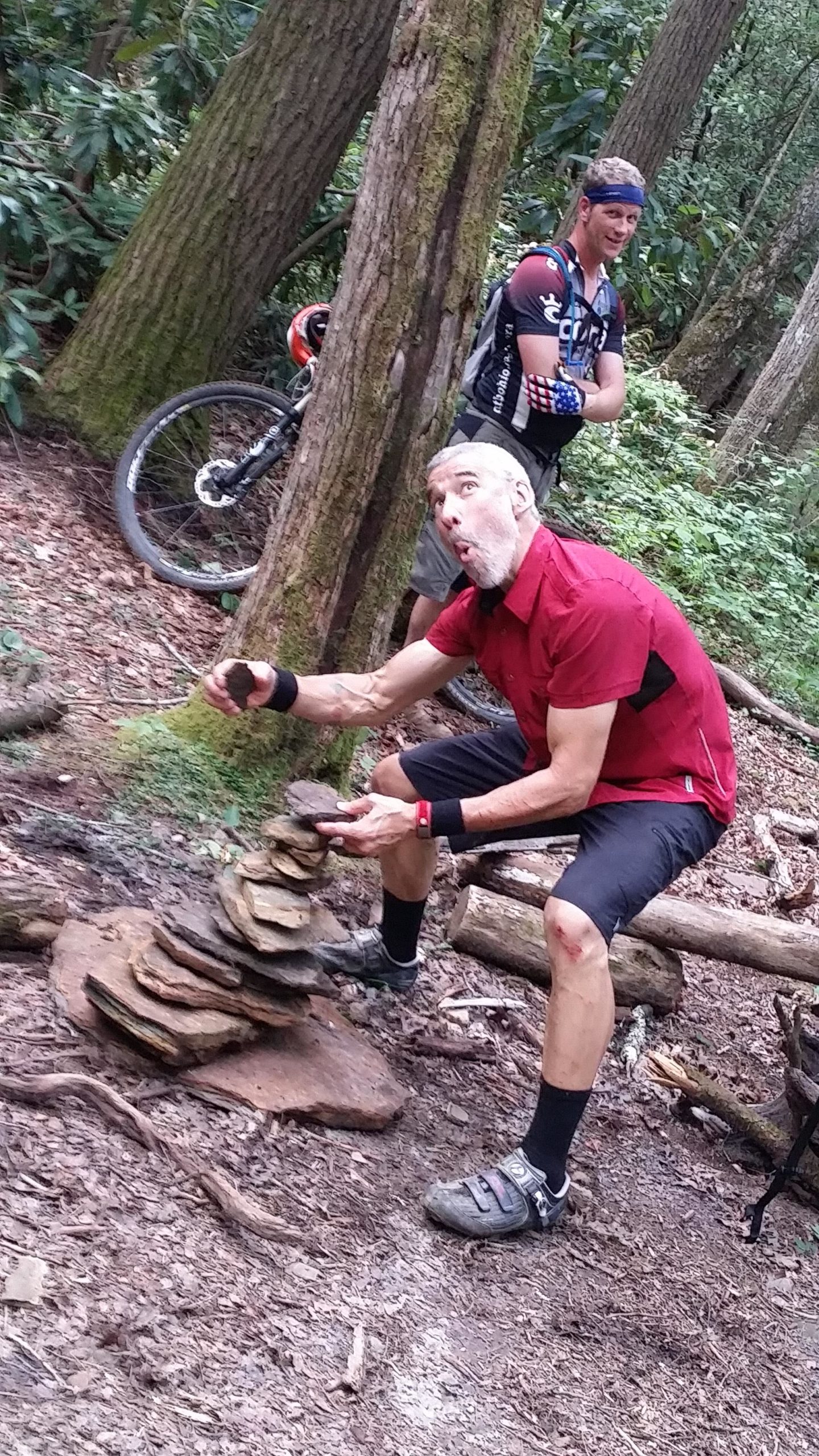 A man in a red shirt is balancing rocks while seated on the forest floor, looking surprised and focused. In the background, another man with a bike and a headband watches, smiling. Surrounding them are trees and greenery typical of a woodland area. The ground is covered with leaves and twigs, indicating a natural hiking or biking trail. DuPont State Forest mountain bike trail.