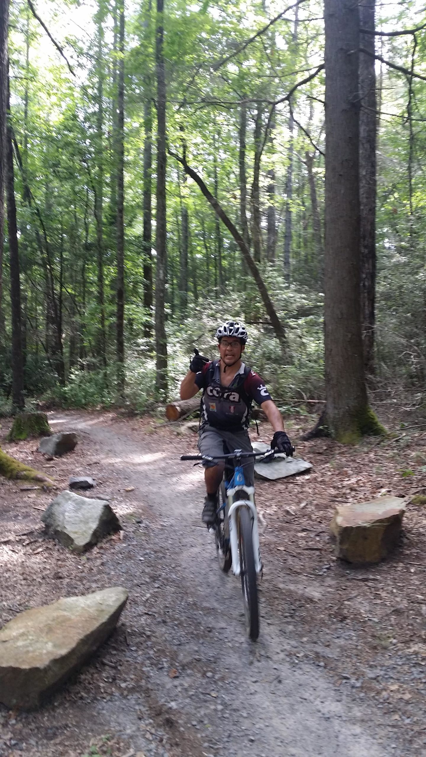 A person on a mountain bike riding along a dirt path through a dense forest, smiling and giving a thumbs-up gesture. Surrounding trees and rocks line the pathway, creating a natural, wooded atmosphere. DuPont State Recreational Forest mountain bike trail.