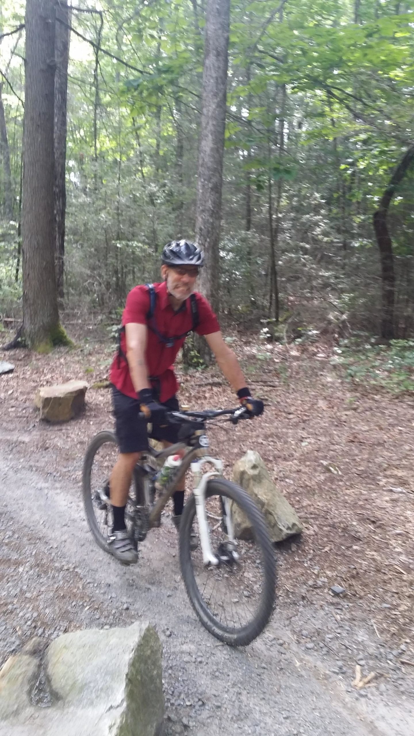 A person in a red shirt and black shorts rides a mountain bike along a wooded trail, surrounded by trees and rocks. The cyclist is wearing a helmet and gloves, and appears to be smiling as they navigate the path. DuPont State Forest mountain bike trail.