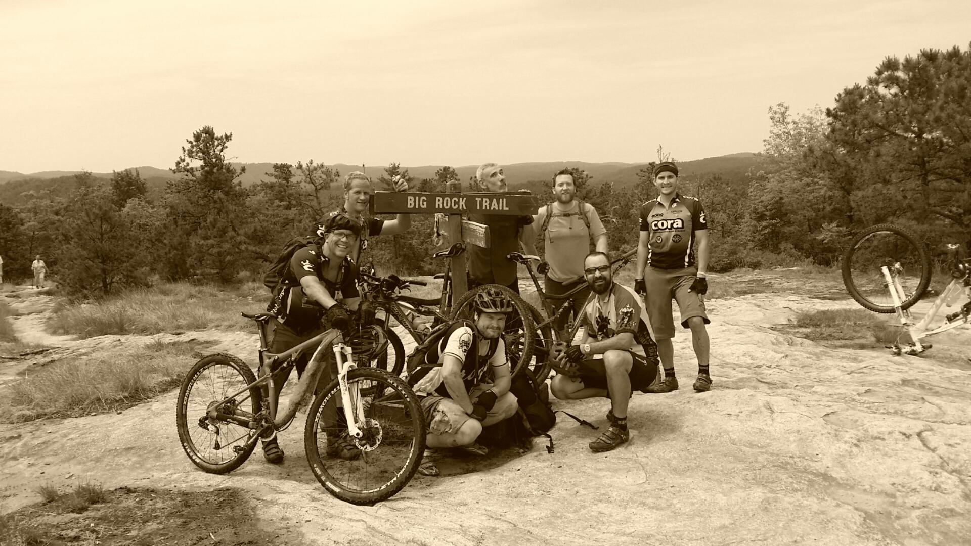A group of six mountain bikers posing for a photo near a trail sign labeled "Big Rock Trail." They are surrounded by a natural landscape of trees and rocky terrain. Some of the bikers are kneeling while others are standing, all smiling and wearing biking gear. In the background, a person can be seen walking on a trail. The image has a sepia tone, giving it a vintage feel. DuPont State Recreational Forest mountain bike trail.