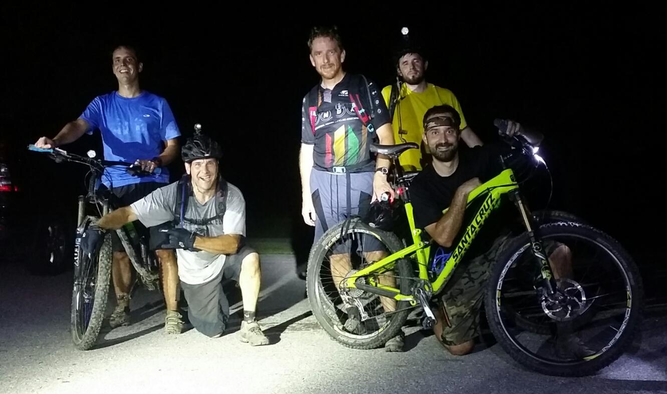 A group of five mountain bikers poses for a photo at night, illuminated by bicycle headlights. Two men are standing on bikes, while three others are squatting beside their bikes. The riders are wearing casual athletic clothing, and their bikes appear equipped for off-road use. The background is dark, suggesting they have just finished a nighttime ride. England Idlewild Mountain Biking Park mountain bike trail.
