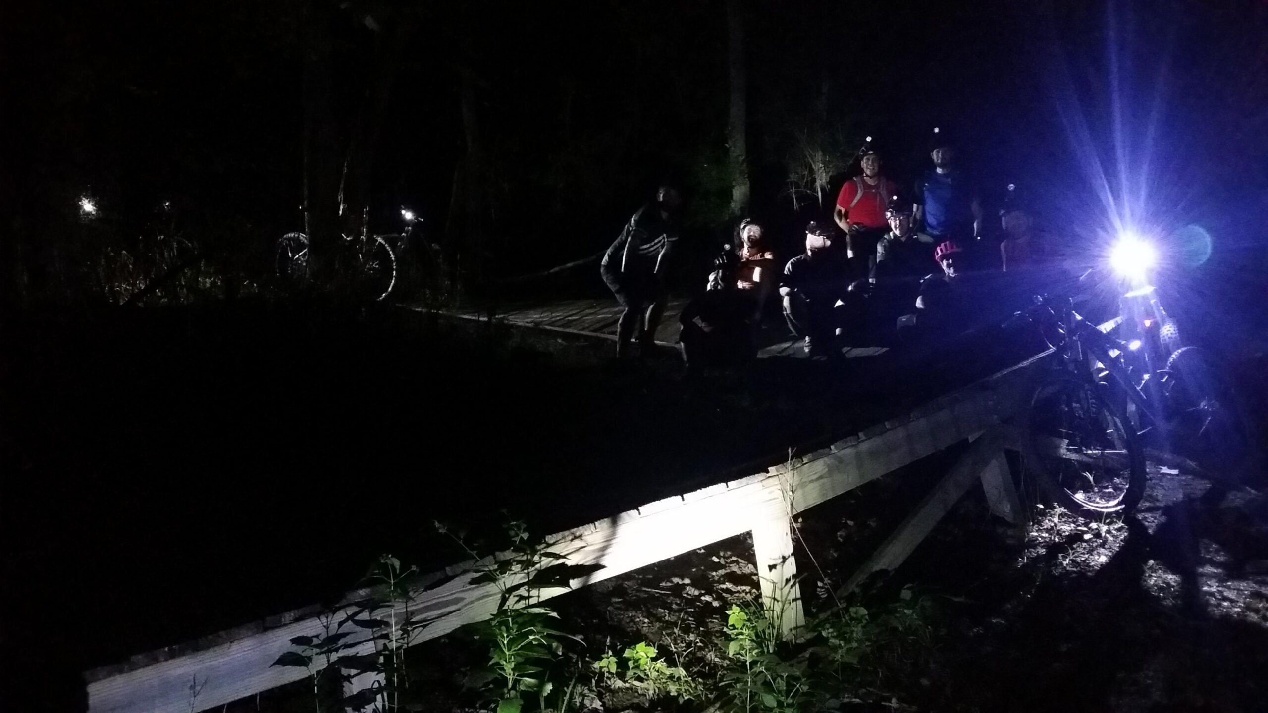 A group of mountain bikers gathered at night on a wooden bridge, illuminated by their bike lights and headlamps. The surrounding area is dark, with only the beams of light highlighting the cyclists and the path ahead. Vegetation can be seen near the edge of the bridge. England Idlewild Mountain Biking Park mountain bike trail.