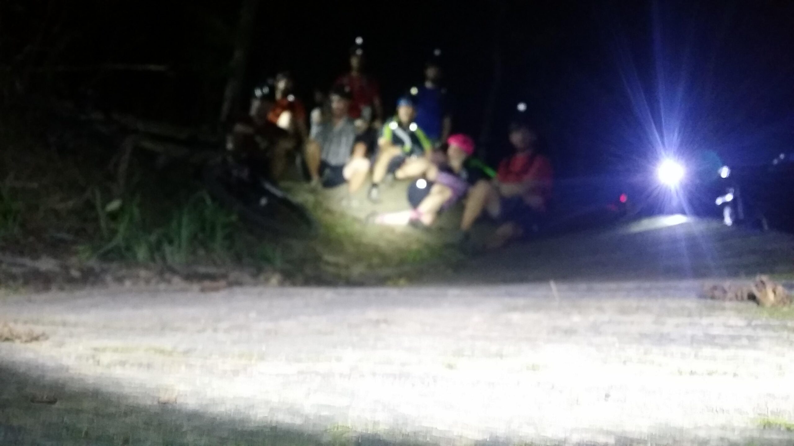 A group of cyclists sitting together on a trail at night, wearing headlamps and illuminated by a distant light. The scene is slightly out of focus, with a gravel path in the foreground and trees in the background. England Idlewild Mountain Biking Park mountain bike trail.