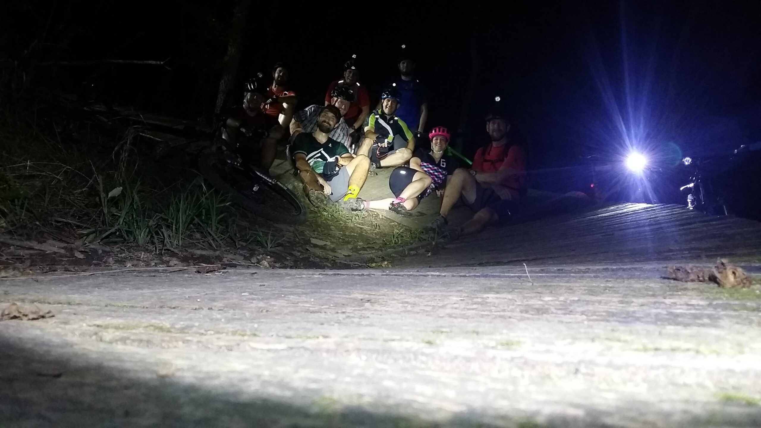 A group of mountain bikers gathered on a path at night, illuminated by their bike lights and headlamps. They are sitting in a relaxed pose, surrounded by trees and nature, with bicycles parked nearby. The scene captures a sense of camaraderie and adventure in a nighttime outdoor setting. England Idlewild Mountain Biking Park mountain bike trail.