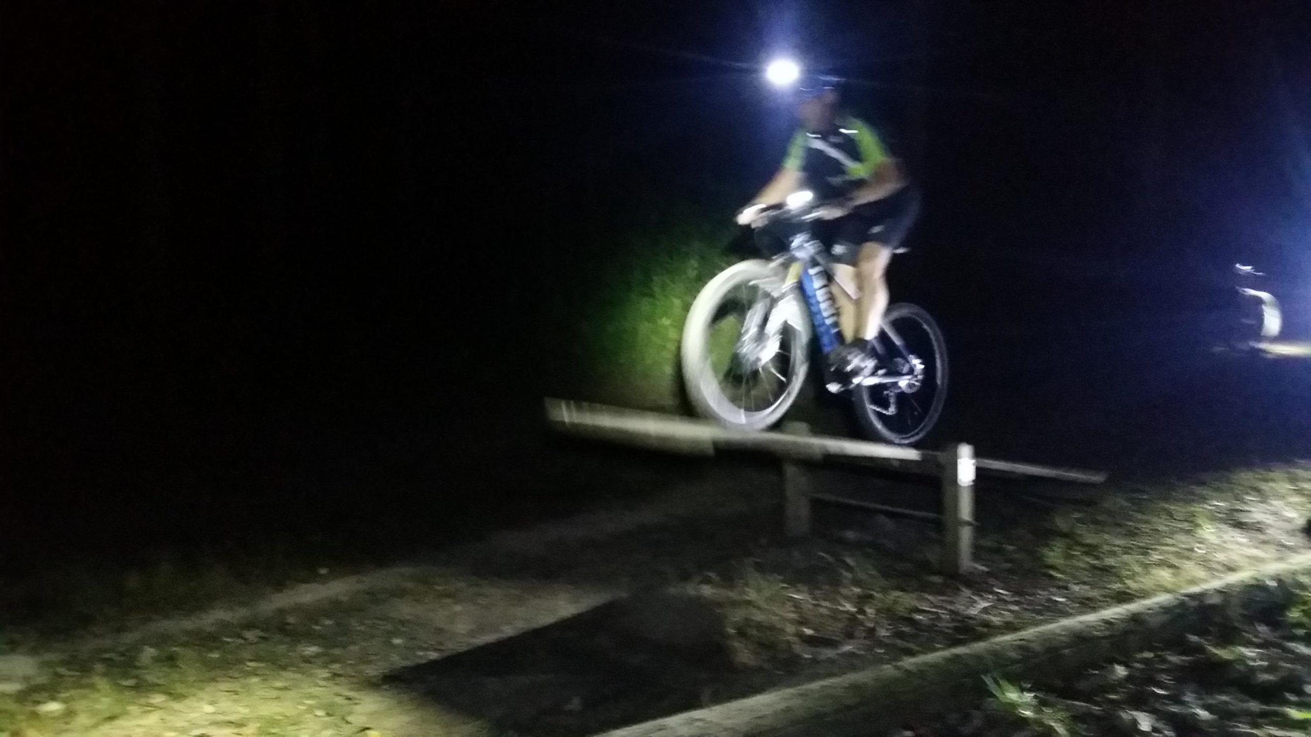 A cyclist wearing a headlamp jumps over a wooden ramp on a dirt trail at night, illuminated by the light from the bike's headlamp and nearby vehicle headlights. England Idlewild Mountain Biking Park mountain bike trail.