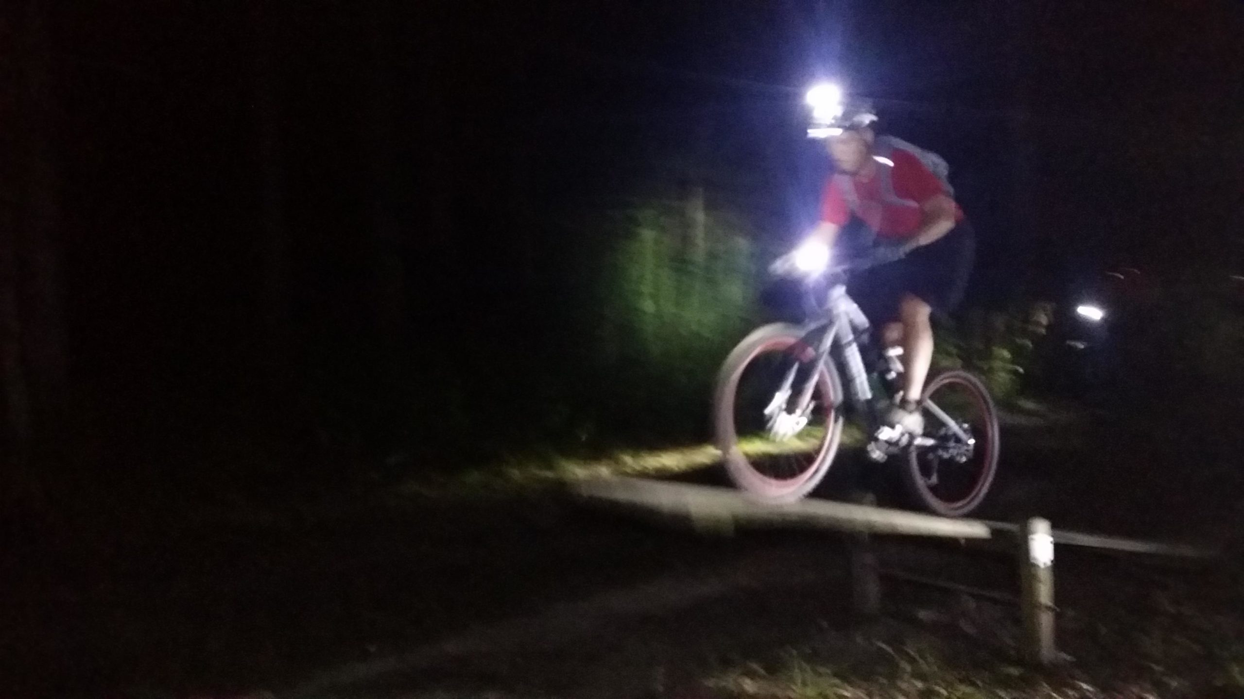 A cyclist wearing a headlamp rides a mountain bike over a wooden ramp at night, with trees and faint light in the background. England Idlewild Mountain Biking Park mountain bike trail.