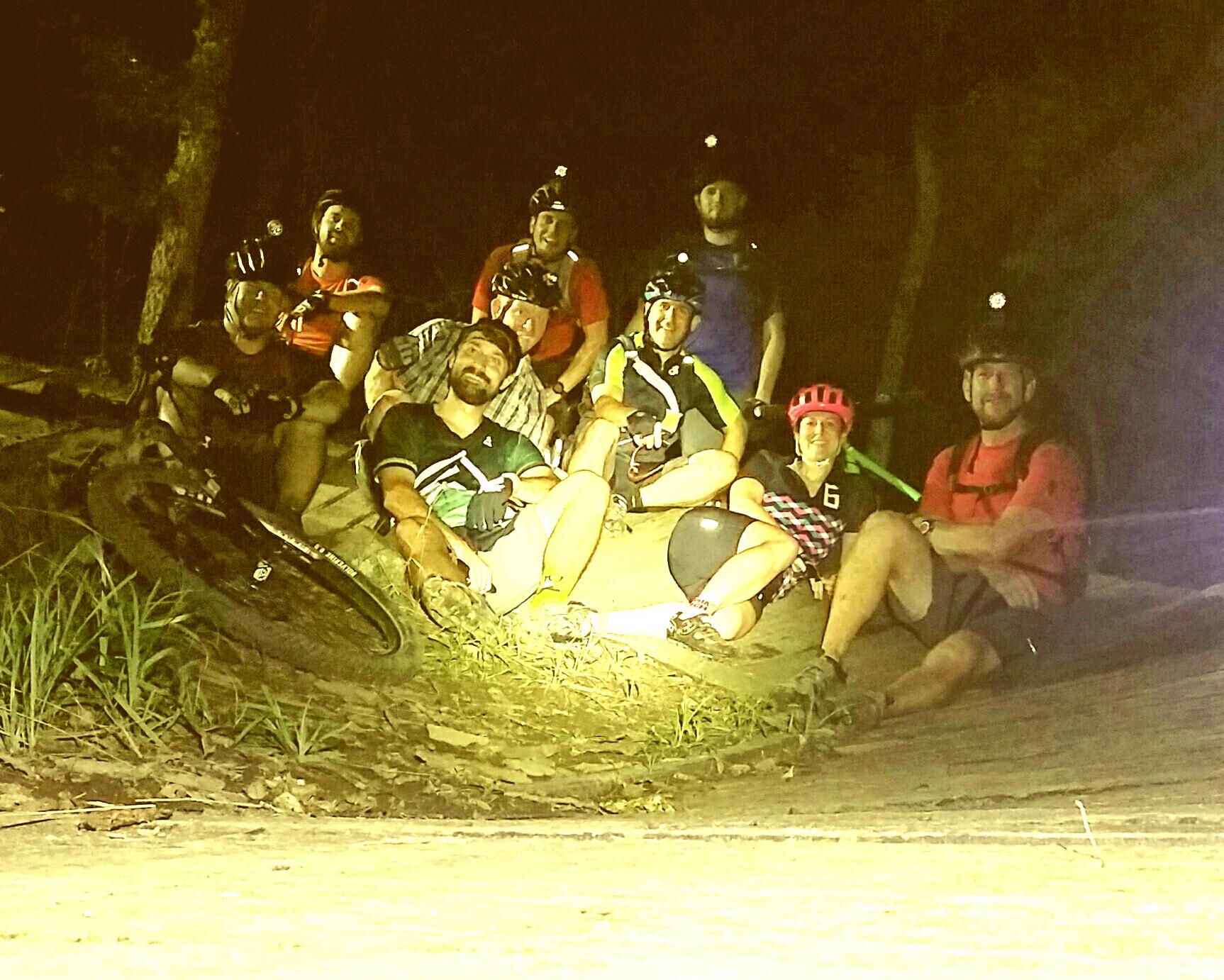 A group of eight mountain bikers sitting together on a rock during a night ride, wearing helmets with headlamps. The setting is dark, surrounded by trees, and they are smiling and enjoying their break. Some riders are in colorful cycling jerseys, and a bicycle is leaning against the rock nearby. England Idlewild Mountain Biking Park mountain bike trail.
