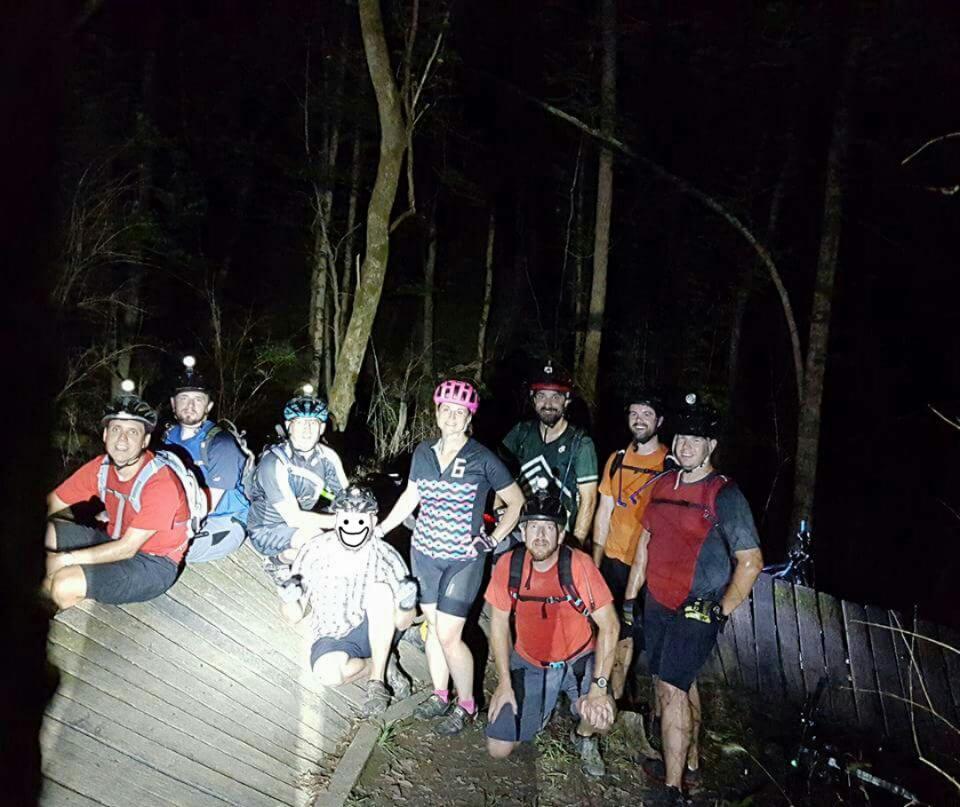 A group of eight mountain bikers poses for a photo at night in a wooded area, wearing helmets with lights. They are gathered around a wooden structure, smiling and enjoying their outdoor adventure. The scene is illuminated by their headlamps, highlighting the trees and the group's camaraderie. England Idlewild Mountain Biking Park mountain bike trail.