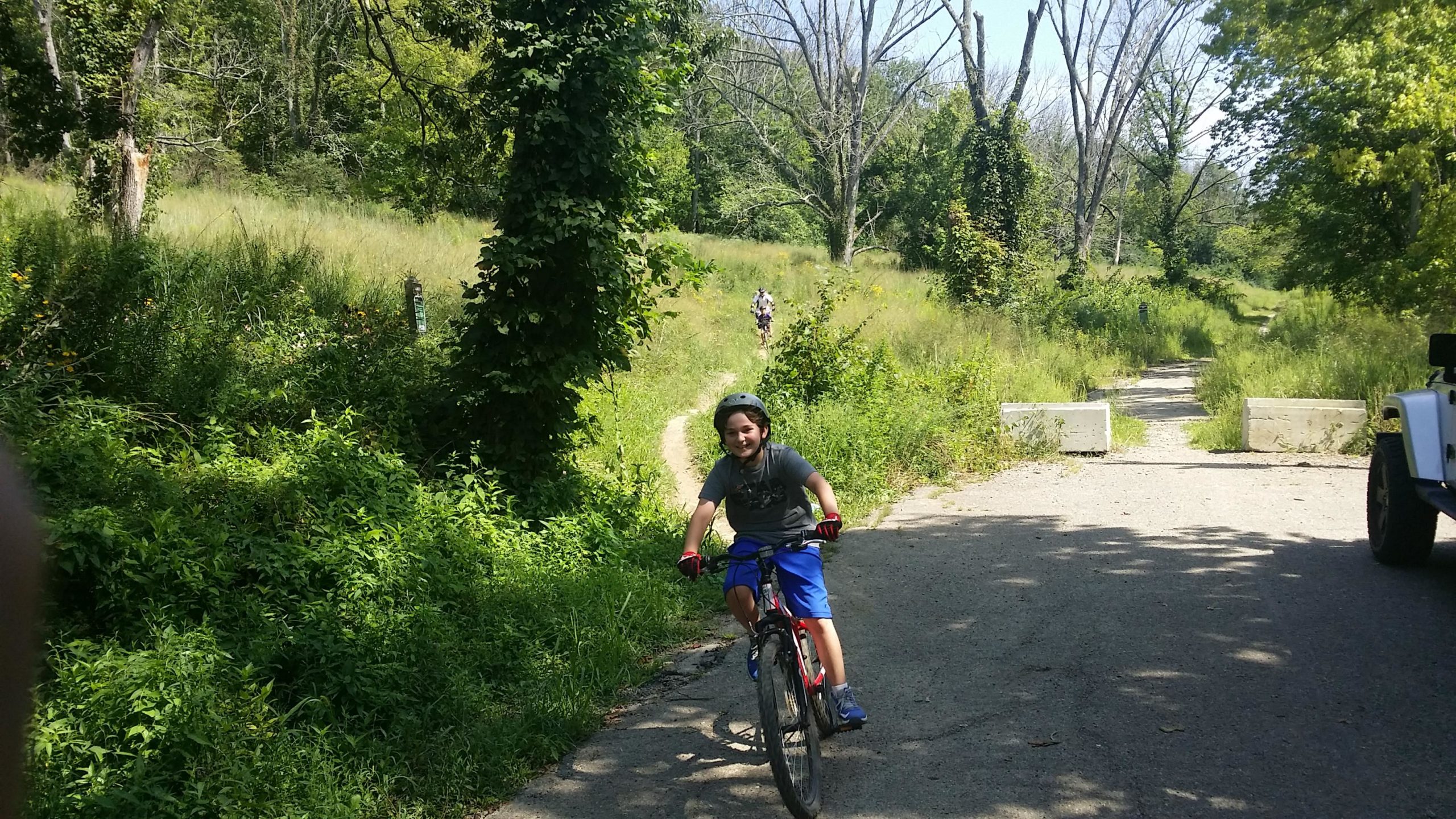 A young boy wearing a helmet rides a bicycle down a dirt path surrounded by lush greenery. In the background, a person can be seen walking along the trail, while concrete barriers mark the end of the path. The scene captures a sunny day in a natural outdoor setting. Devou Park mountain bike trail.