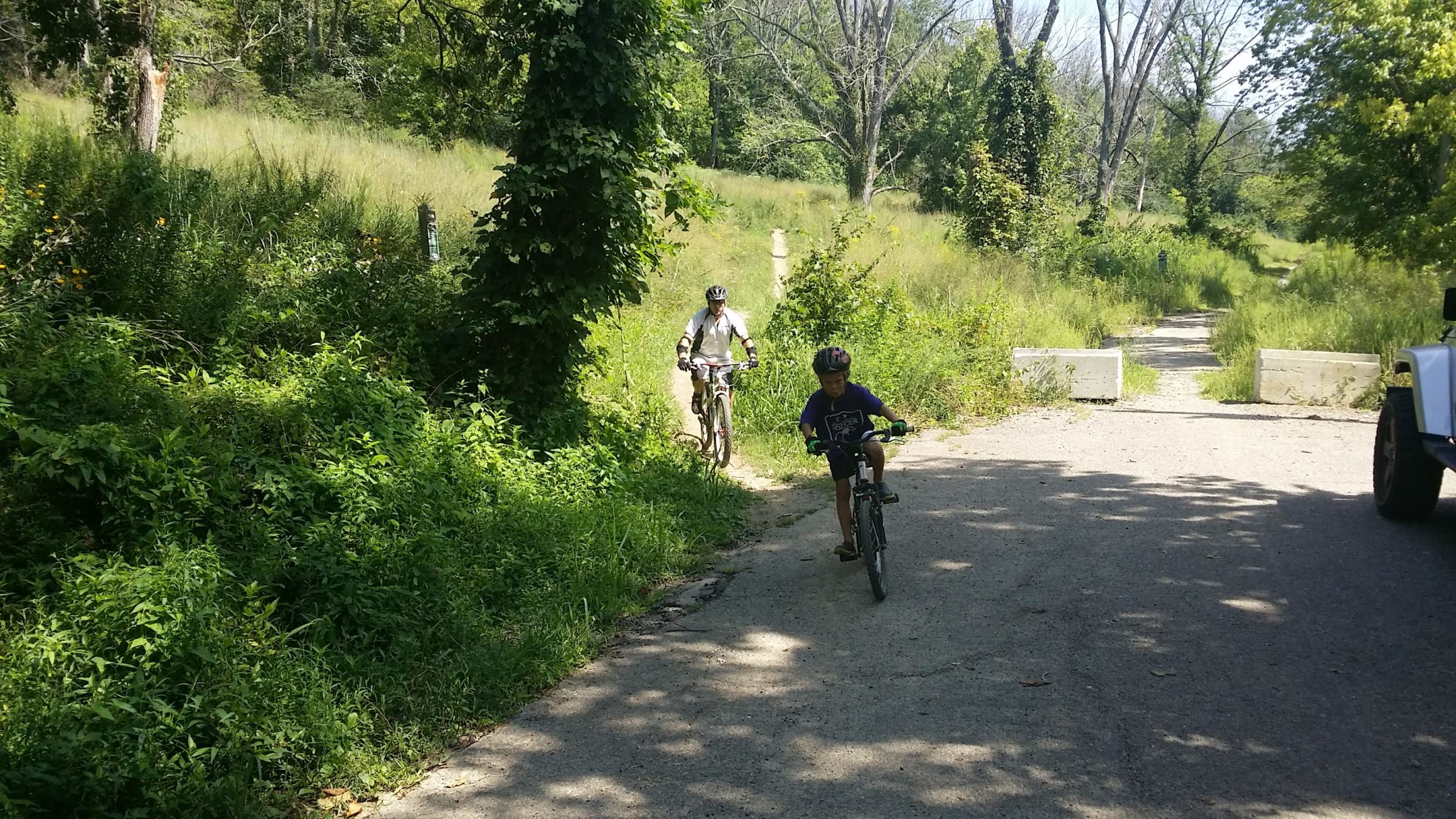 Two individuals riding bicycles on a gravel path surrounded by lush greenery and tall grass. One is a child in a purple shirt riding ahead, while an adult follows closely behind. The scene is set in a sunny environment, with trees and a barrier in the distance. Devou Park mountain bike trail.