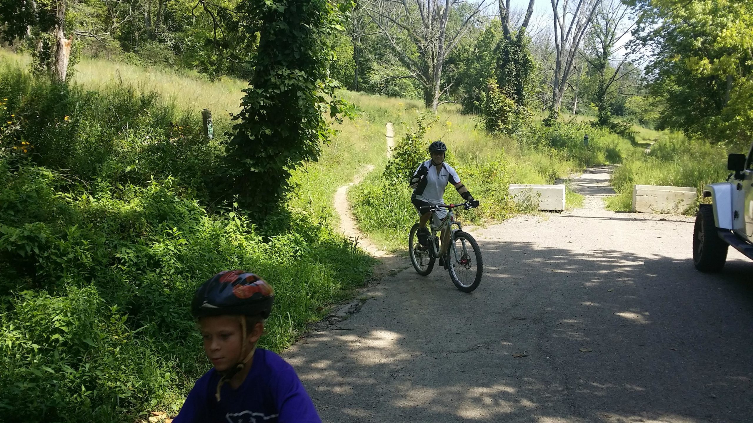 Two children are riding bicycles along a scenic path surrounded by lush greenery. One child is pedaling on the road, while another is in the foreground, focusing ahead. The trail leads to a grassy area with sparse trees and vegetation in the background, indicating a peaceful outdoor environment. Devou Park mountain bike trail.