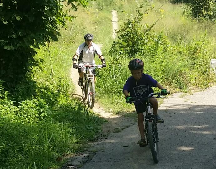A young boy riding a bicycle on a gravel path, with a man on a mountain bike following behind him. Both are surrounded by lush greenery and tall grass on a sunny day. Devou Park mountain bike trail.