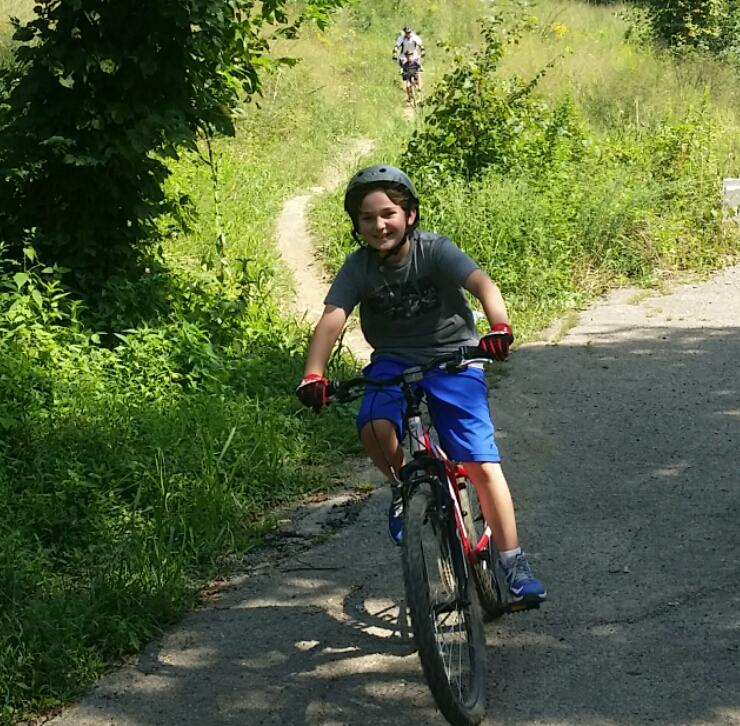 A young boy riding a bicycle on a dirt path surrounded by greenery, smiling and wearing a helmet. In the background, another cyclist is riding along the same path. The scene captures a sunny day outdoors, showcasing an enjoyable recreational activity. Devou Park mountain bike trail.