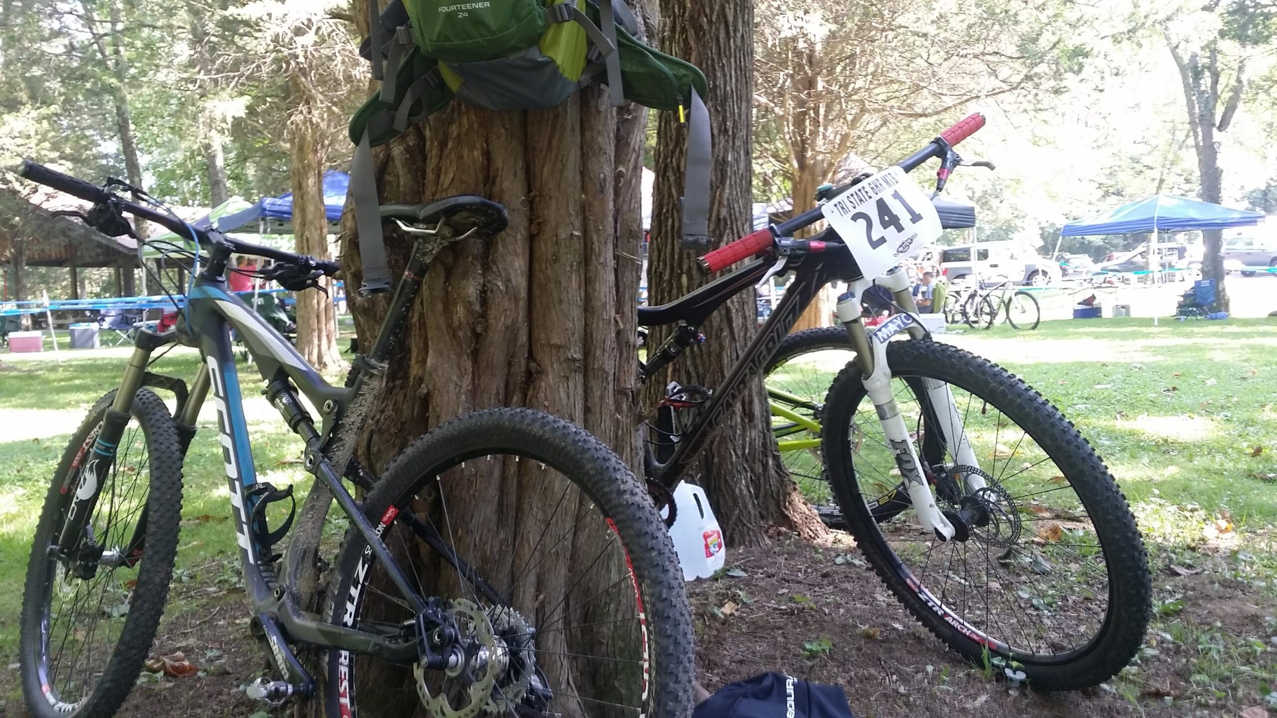 Two mountain bikes are leaning against a tree in a green park setting. One bike is gray with blue accents and has a prominent water bottle holder, while the other is black with a white front fork and a race number 241 attached to the handlebars. In the background, tents and other bicycles are visible, indicating an outdoor event. A backpack hangs from the tree above the bikes. Versailles State Park mountain bike trail.