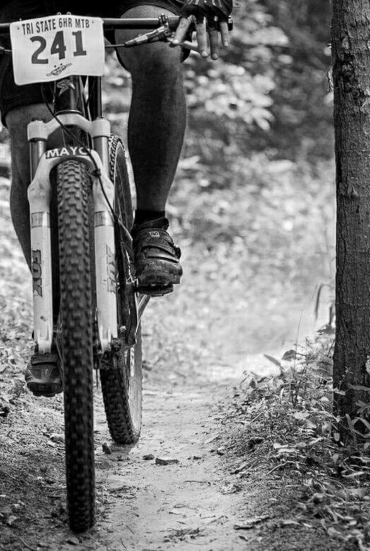 A close-up black and white photograph of a mountain biker's lower half, showing one foot on the pedal and the bike's tire on a narrow dirt trail. The bike has a race number plate labeled "241," and the background features blurred trees and foliage, suggesting a forested environment. England Idlewild Mountain Biking Park mountain bike trail.
