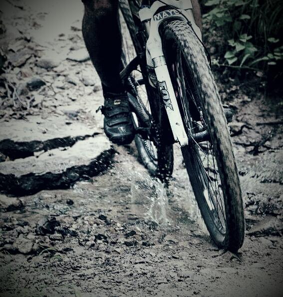 Close-up of a mountain bike wheel splashing through a muddy section of a trail, with a cyclist's foot on the pedal. The background features rocky terrain and greenery. England Idlewild Mountain Biking Park mountain bike trail.
