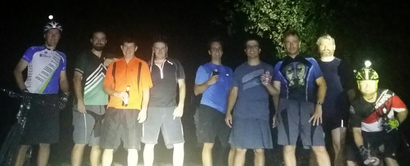 A group of nine male cyclists poses for a photo at night, surrounded by trees. Some are holding drinks, and the cyclists are dressed in a variety of athletic clothing and gear, with one wearing a helmet equipped with a light. Light from a nearby source illuminates the group. England Idlewild Mountain Biking Park mountain bike trail.